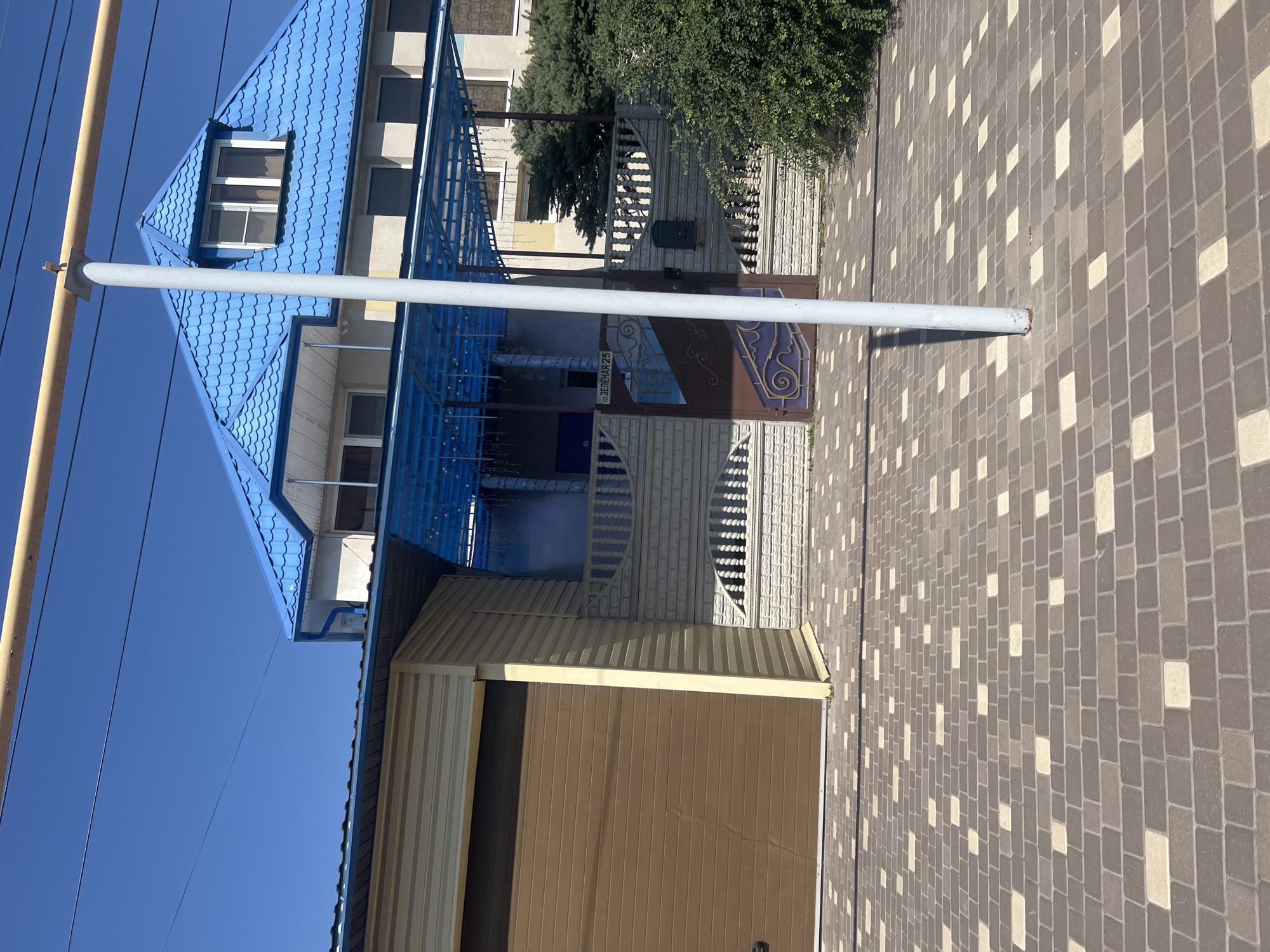 The image shows a residential property on a sunny day. The scene features a two-story house with a distinctive blue, wavy metal roof. To the left of the house is a beige siding garage with a brown rolling door. A light-colored fence with decorative white balusters and a dark brown gate with blue and purple accents stands in front of the house. A street sign on the gate reads "ул. ЗЕЛЕНАЯ 25". The ground in front of the property is paved with a pattern of brown and light tan rectangular pavers. A grey metal pole, possibly a light post, is positioned in the foreground, casting a shadow on the pavement. Dense green foliage is visible to the right of the gate. Power lines are strung across the top of the frame against a clear blue sky. This property is located in Kilyakovka, Russia.