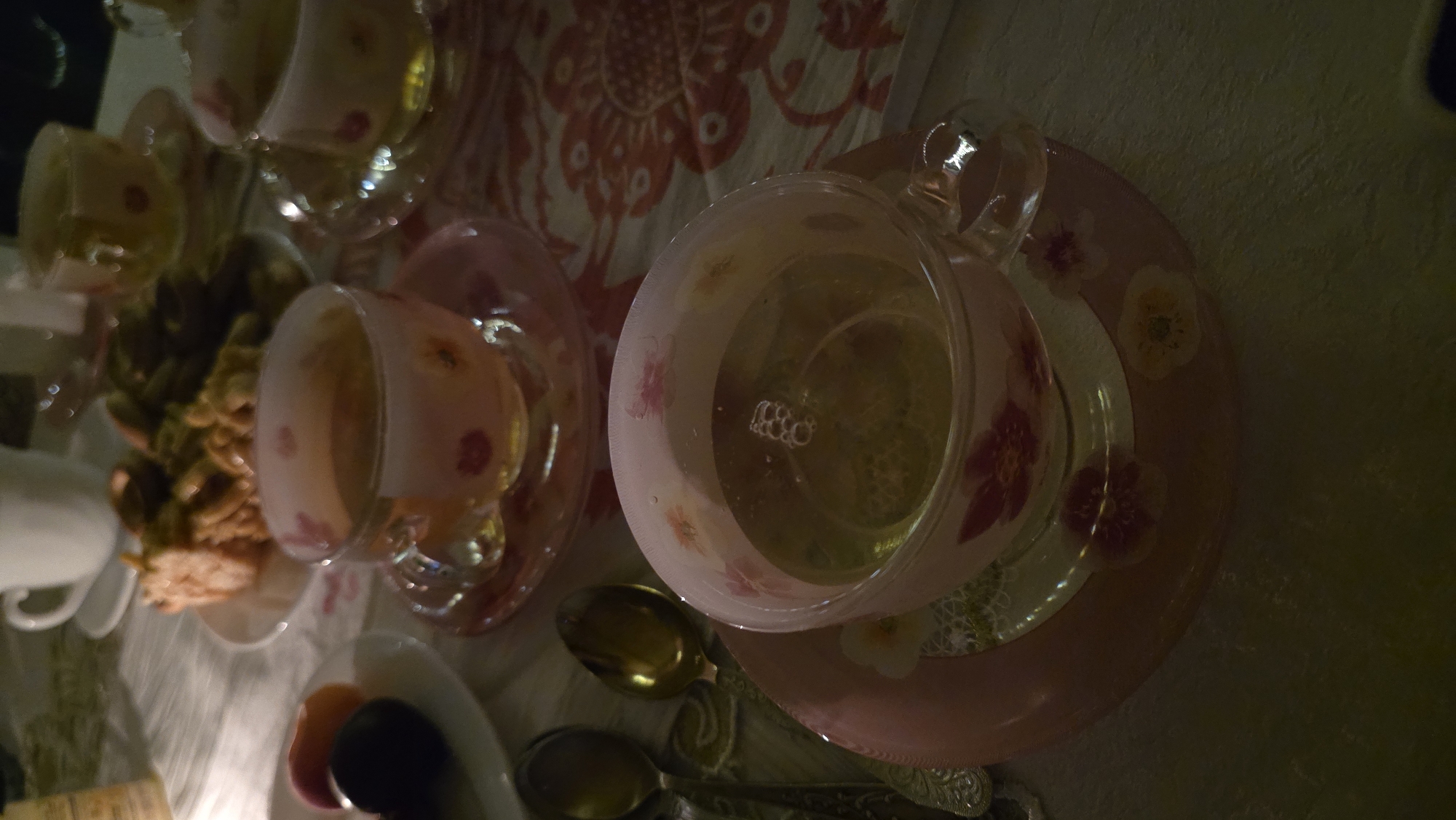 The image depicts a close-up, overhead view of a tea set on a table. In the foreground, a clear glass teacup with a pink floral pattern rests on a matching pink saucer. The teacup is filled with a light-colored liquid, likely tea, with small bubbles visible on the surface. To the left of the teacup, two antique silver spoons are partially visible. In the background, another similar teacup and saucer are present. Behind the teacups, a plate holds a portion of what appears to be a baked good, possibly a cake or pastry, with a stack of green items, possibly vegetables or candies, next to it. A white ceramic mug is also visible in the far background. The table is covered with a white tablecloth patterned with red floral motifs. The lighting is dim, suggesting an indoor setting at dusk or night.