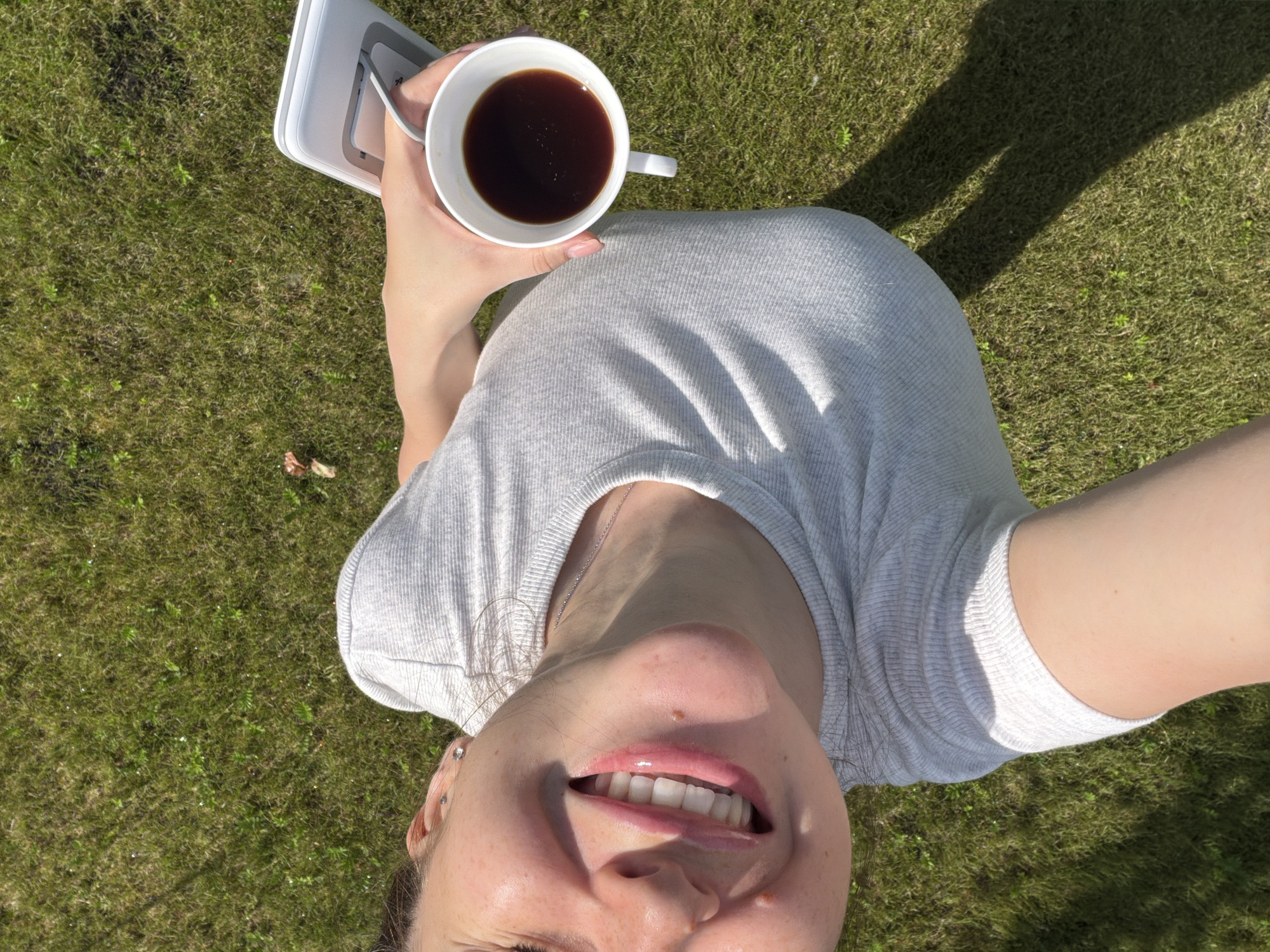 In Miasskoye, Russia, a person is captured from a top-down perspective, holding a white coffee cup filled with dark liquid in their right hand. The person is wearing a light gray, ribbed t-shirt and is positioned on a patch of green grass. Their face is turned towards the camera, with a wide, beaming smile showcasing their teeth. The sunlight casts a noticeable shadow across the grassy area. Behind the person, a white electronic device with a stand is visible. The environment appears to be outdoors on a sunny day.