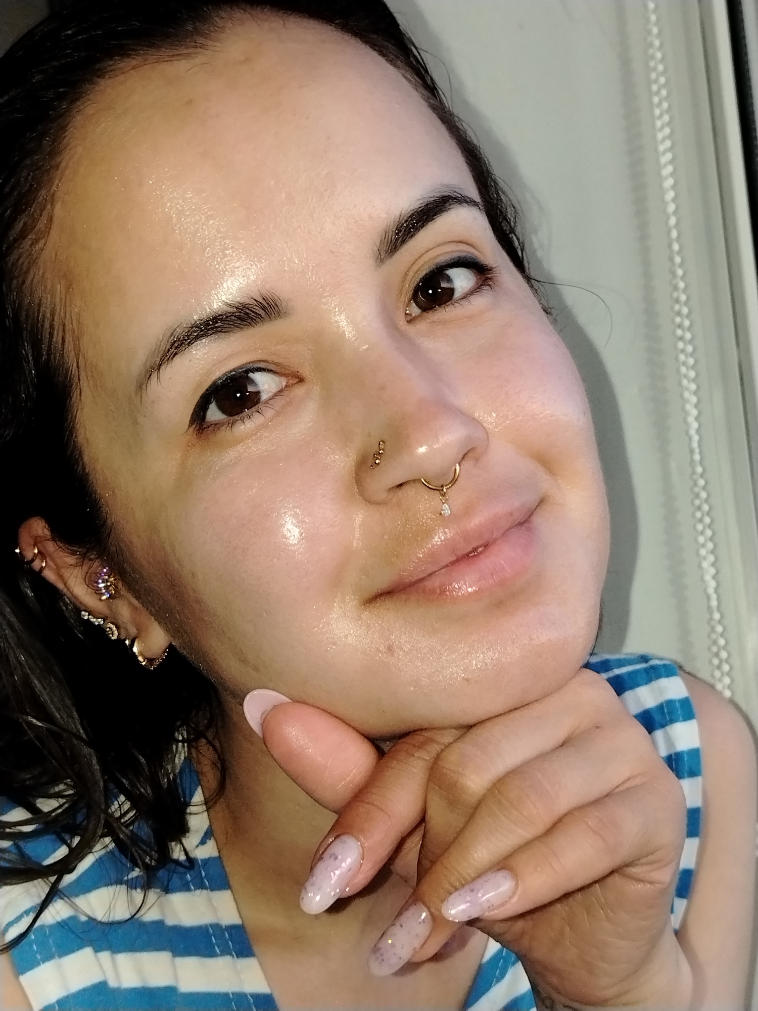 A close-up of a woman's face and hand, with window blinds visible in the background. The woman has brown hair, brown eyes, and fair skin. She has multiple piercings in her left ear and nose. Her fingernails are light pink and almond-shaped. She is wearing a blue and white striped shirt. Her hand is positioned with her fingers touching her chin. The photo does not provide information regarding the woman's location.