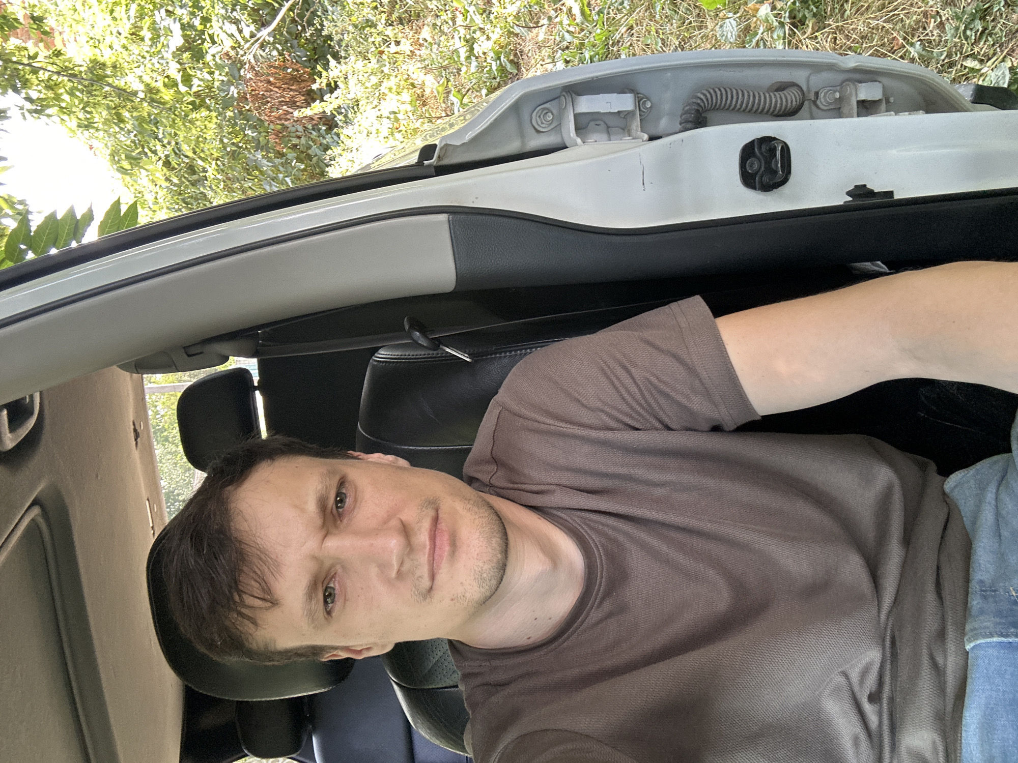 A man is sitting in the driver's seat of a white car, with the driver's side door open. He is wearing a brown t-shirt and blue jeans. He has brown hair and a light complexion. His expression is neutral. The car is parked on a dirt road, with trees and foliage visible in the background. The scene appears to be in a rural area, possibly on the outskirts of Makiv, Ukraine.