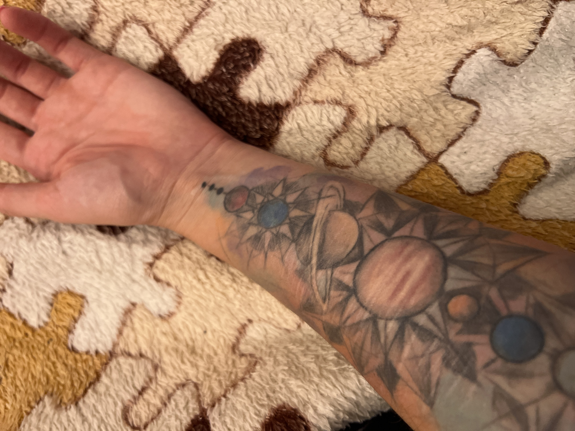 The image shows a person's forearm with a tattoo of planets and geometric shapes. The tattoo is located on the underside of the forearm, extending from the wrist upwards. The planets are depicted in various colors, including red, blue, and orange, and are surrounded by star-like patterns and abstract designs. The background of the image is a textured blanket with a pattern of interlocking shapes in shades of brown, beige, and white. The lighting is soft, and the image is taken from a slightly elevated angle. There is no indication of the location in Krasnoe-na-Volge, Russia.