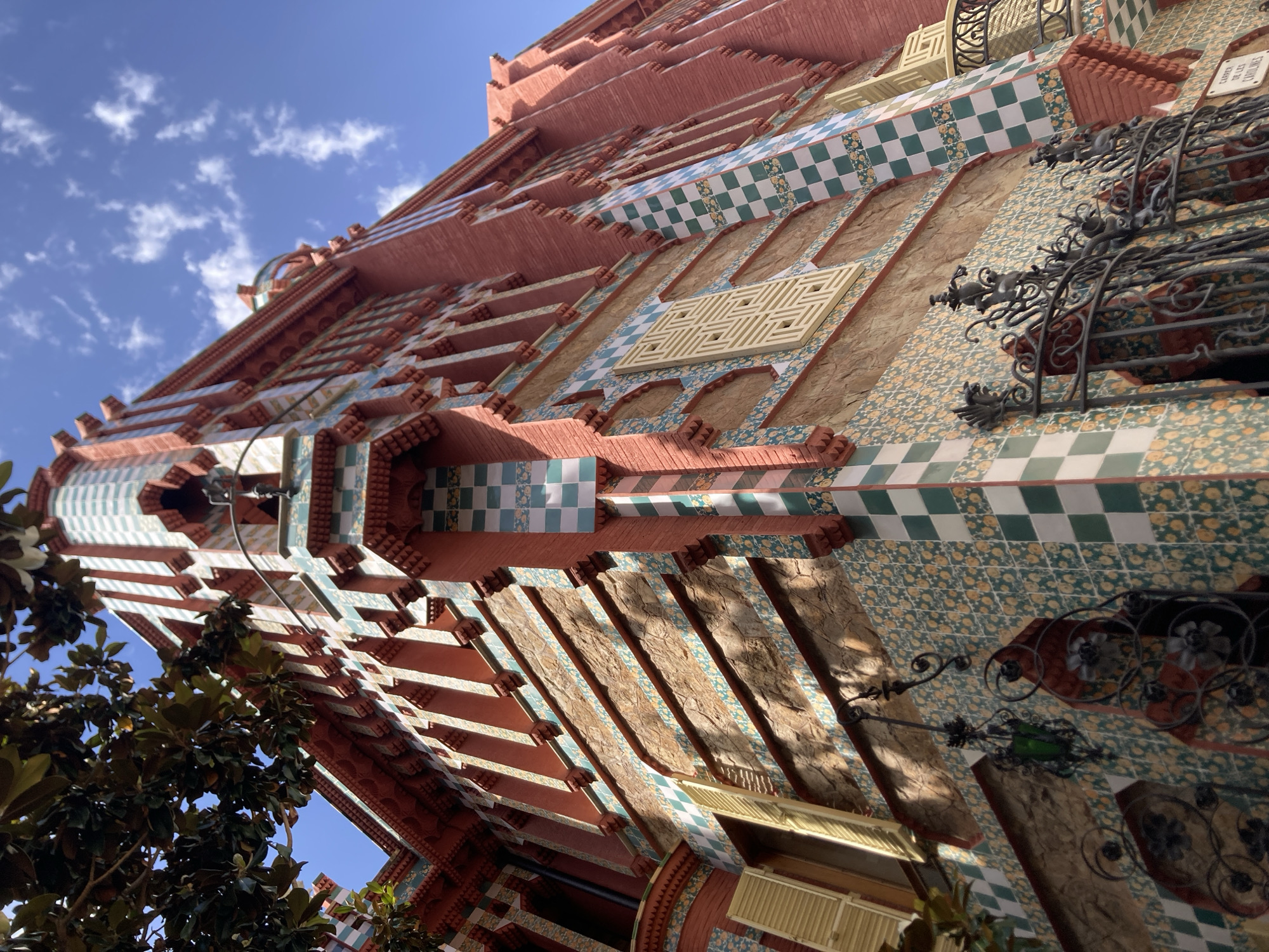 This is a low-angle, full-shot photograph of the exterior of Casa Vicens, located in the Gràcia district of Barcelona, Spain. The building exhibits a vibrant and eclectic architectural style, characterized by its red brick facade adorned with colorful ceramic tiles. These tiles feature intricate geometric patterns, checkerboard designs in green and white, and floral motifs in yellow and green. The building's upper sections are decorated with red brickwork and contrasting turquoise and white checkered tiles.
Several architectural elements are visible, including arched windows with red decorative framing, a small balcony with a wrought-iron railing, and elaborate wrought-iron gates at the bottom of the frame. A single green glass lantern hangs from a decorative wrought-iron bracket on the wall. The sunlight casts strong shadows across the textured stone portions of the building's lower facade, highlighting the details of the stonework and the tiled surfaces. A portion of a leafy tree branches in from the upper left corner of the image, providing a natural contrast to the man-made structures. The sky above is a clear blue with scattered white clouds. There are no people visible in the image.