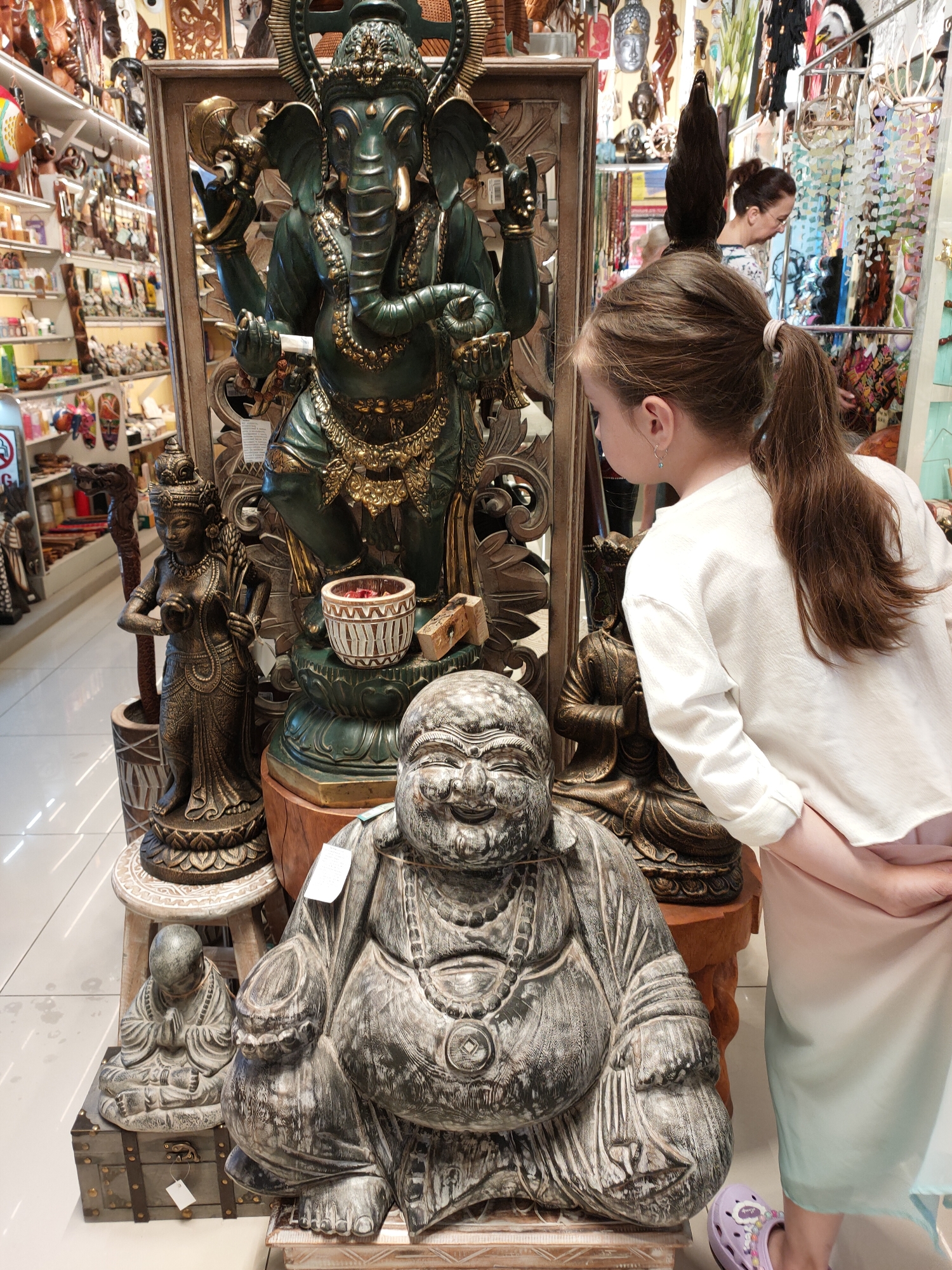 In a retail store in Sochi, Russia, a young girl with brown hair tied in a ponytail stands facing a display of statues. She is wearing a long-sleeved white shirt and a light-colored skirt, with a purple sandal on her left foot. To her left, a large, dark green Ganesha statue is prominently displayed, adorned with gold jewelry and holding various objects. In front of the Ganesha statue, a smaller, detailed statue of a woman holding stalks of grain stands on a platform. Further in the foreground, a large, weathered stone statue of a smiling, rotund Buddha is positioned to the right, with a smaller, seated Buddha statue on a wooden chest to its left. In the background, shelves are filled with various decorative items, including masks and small statues. Two other individuals are partially visible in the background, one of whom appears to be an adult woman looking at merchandise. The store appears to be a souvenir or gift shop, with numerous items for sale displayed throughout the space.
