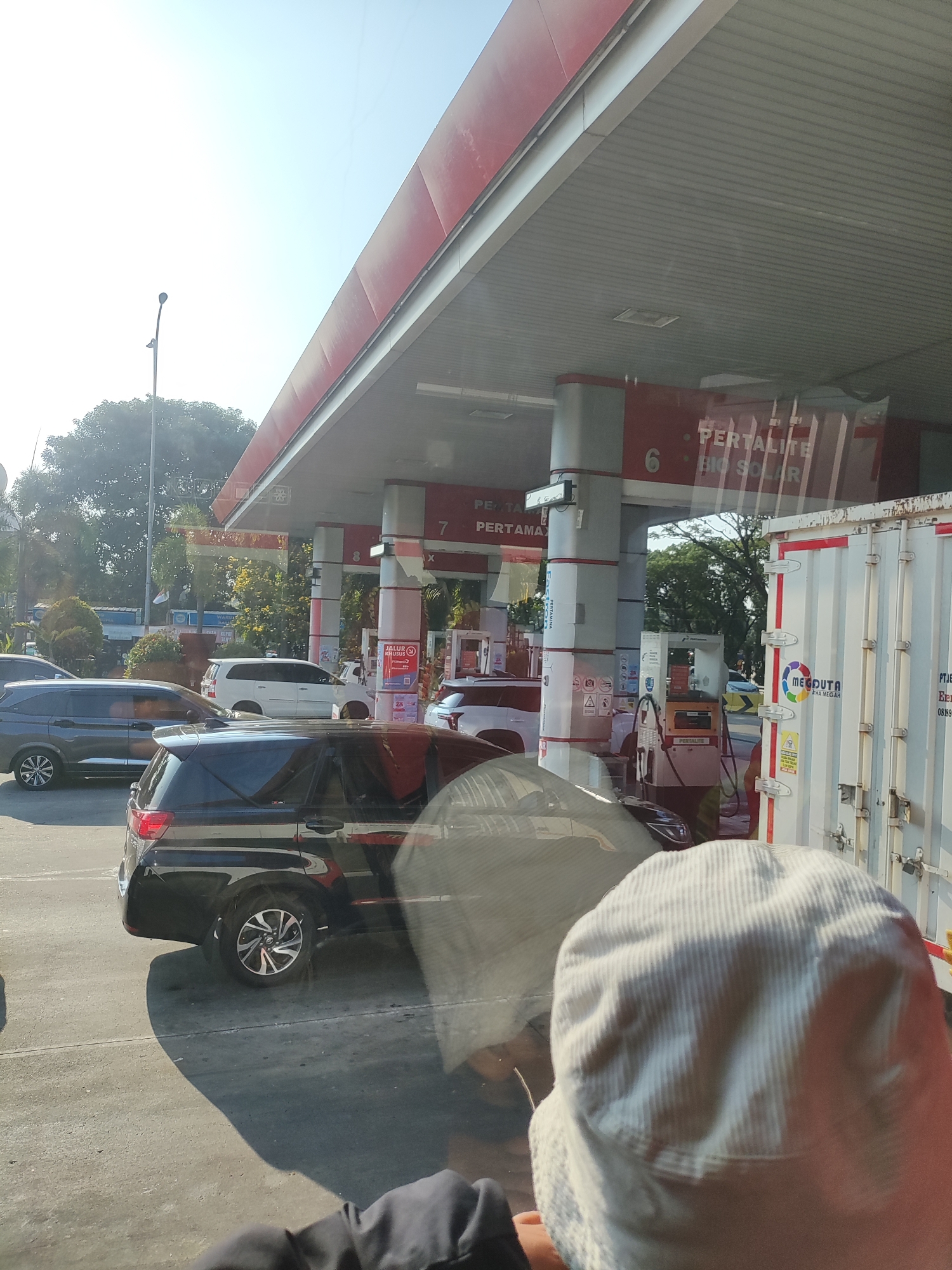 The image captures a scene at a Pertamina gas station in Karawang, Indonesia. The perspective is from inside a vehicle, looking out through the windshield. In the foreground, a person wearing a white bucket hat is partially visible, their head and shoulders obscuring part of the view. Several vehicles are queuing at the gas station. A black SUV is prominently featured in the middle ground, parked at one of the pumps. To its left, a dark gray SUV is also waiting. Further back, a white minivan is visible. The gas station canopy, with its red and white structure, covers the pumps. Signs indicating "PERTALITE" and "BIO SOLAR" are visible on the station's columns. A white truck with "MEDOUTTA" branding is parked to the right. The environment appears to be a sunny day, with trees and a clear sky in the background. The overall impression is a typical busy moment at a roadside service station in an Indonesian city.