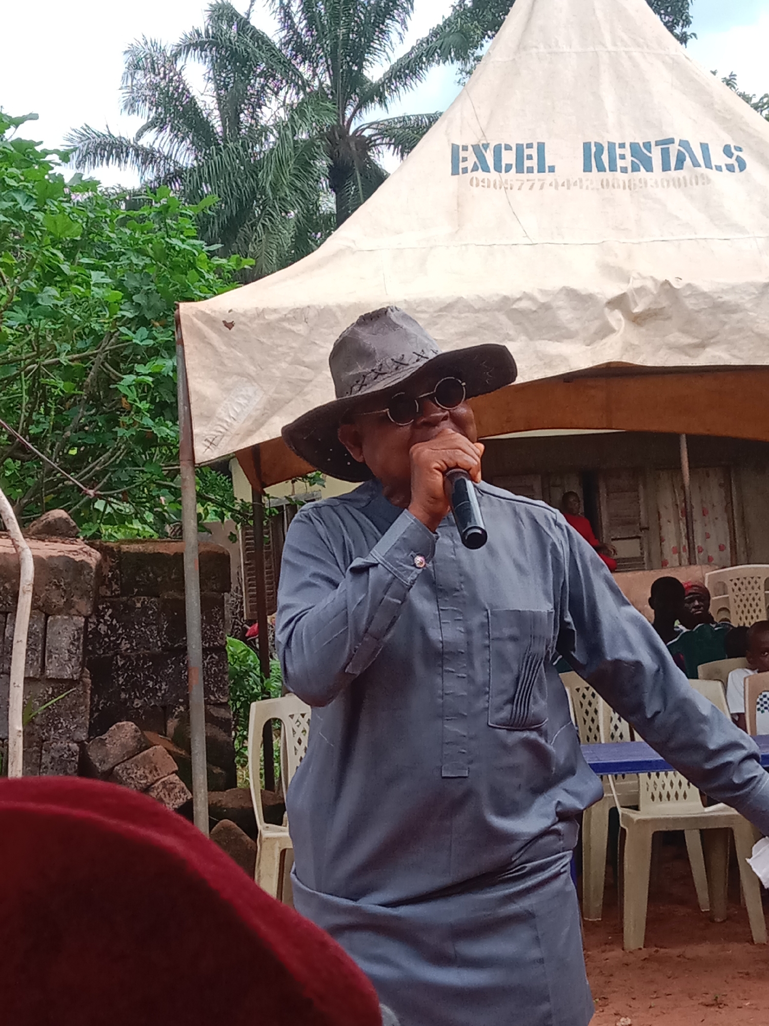An elderly man in Udi, Nigeria, speaks into a microphone while standing under a tent labeled "Excel Rentals." He is wearing a gray suit, a cowboy hat, and sunglasses. People sit in white plastic chairs behind him. There is a brick wall and foliage in the background.