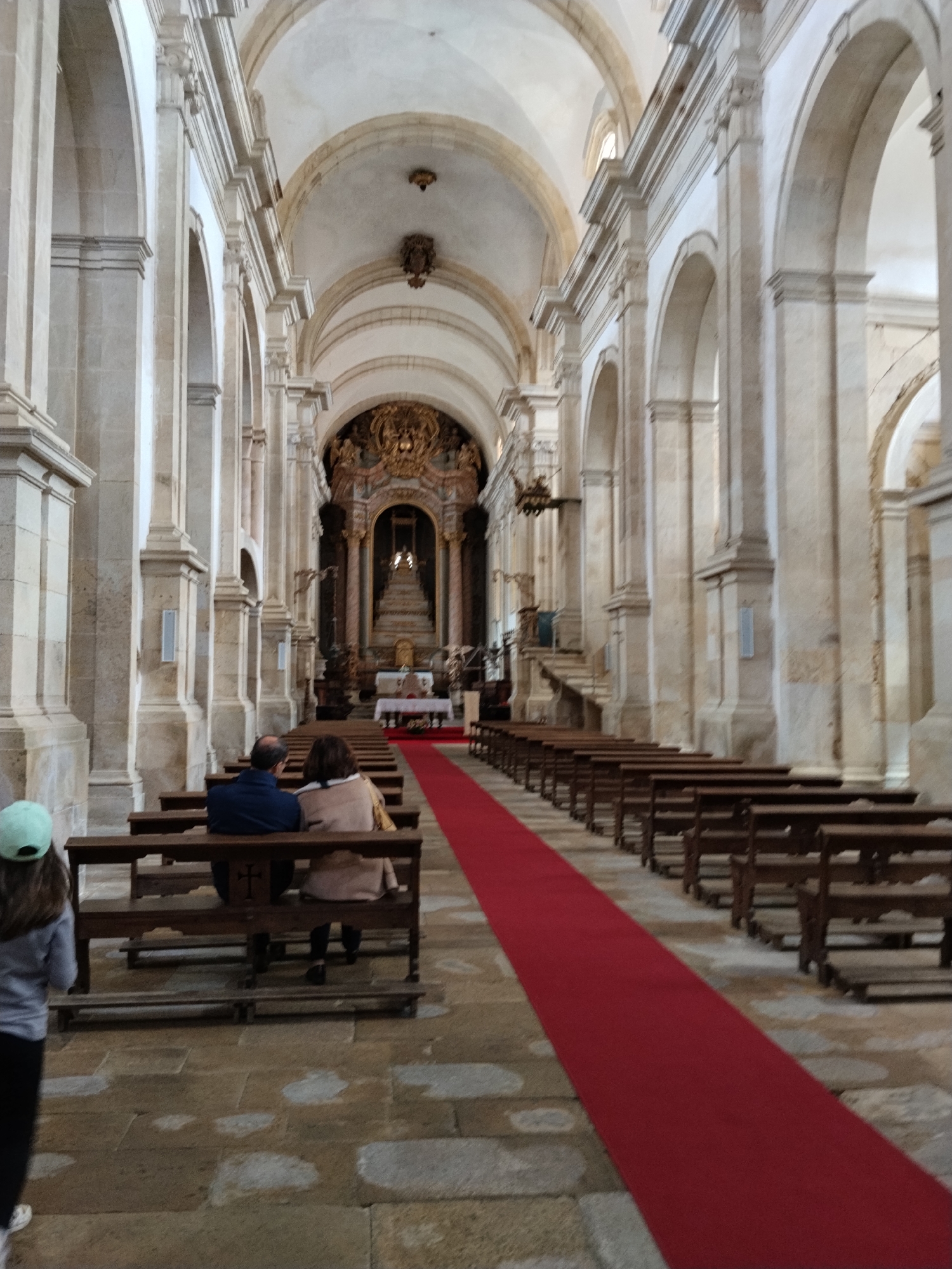 The image depicts the interior of the Mosteiro de Santa Maria de Salzedas in Tarouca, Portugal. A long nave is defined by rows of stone arches and columns, leading to an ornate altar at the far end. A red carpet runs down the center aisle between rows of wooden pews. A couple sits in the front pew, facing the altar. A third person is partially visible in the lower left corner, wearing a light-colored hat. The floor is made of stone tiles.