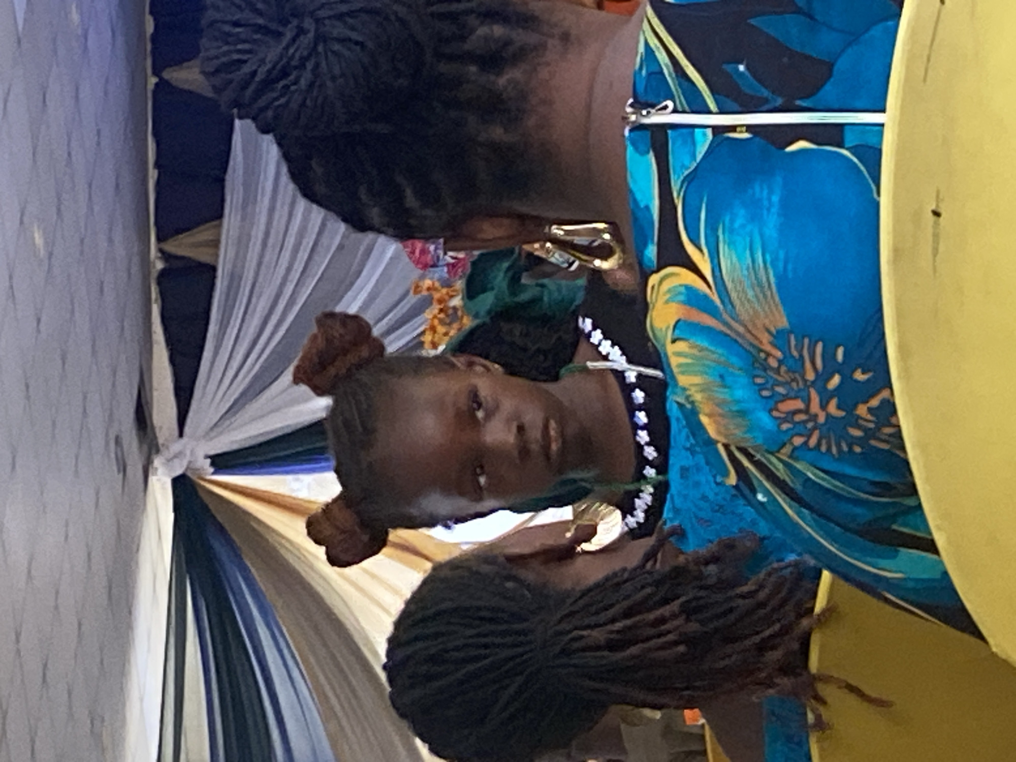 A girl with hair styled in two buns sits flanked by two adults with braided hairstyles. The girl wears a blue lace top with white accents. One adult wears a blue, green, and black floral print top. The setting is decorated with draped fabric in white, tan, and blue hues. The specific location in Gura Top, Nigeria cannot be determined from the image.