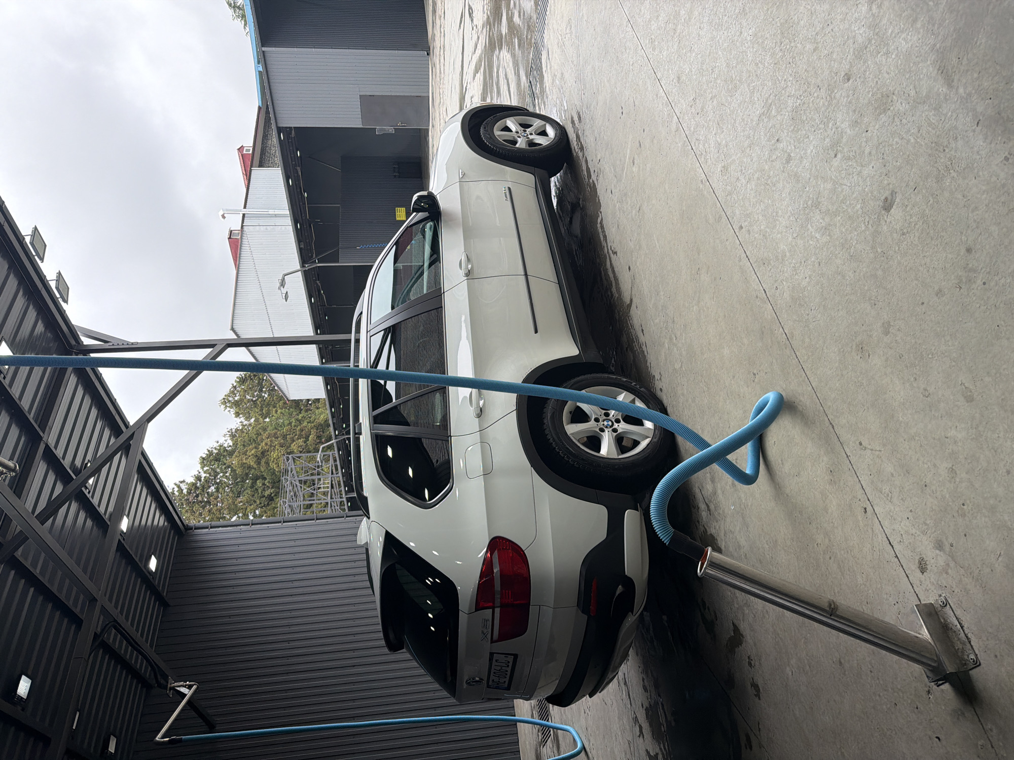 The image shows a white BMW X5 parked at what appears to be a car wash facility, likely in Tsinsvla, Georgia. The car is positioned at an angle on a concrete surface that shows signs of being wet. A blue hose, likely for washing or vacuuming the car, is connected to a metal arm mounted on the ground. The car's license plate is partially visible. The surrounding environment includes corrugated metal walls and roofing typical of a car wash structure, with some vegetation visible in the background. The sky is overcast.