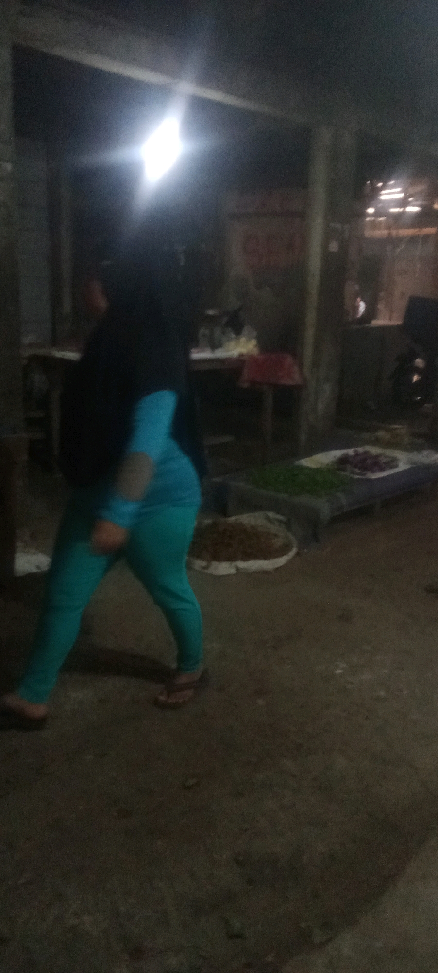 The image depicts an outdoor scene at what appears to be a market or a roadside stall, possibly in Karawang, Indonesia, judging by the context. A person wearing a dark hijab and a turquoise long-sleeved shirt and pants is walking from left to right across the frame. The person is wearing brown flip-flops. In the background, there are various objects, including tables with what seem to be goods for sale, such as produce like green leafy vegetables and red onions. There's also a large, flat surface covered with what looks like a white tarp or cloth, on which the produce is displayed. The environment is dimly lit, with a bright light source visible in the upper left quadrant, casting a glare. The ground appears to be a dirt or unpaved surface. The overall impression is that of a bustling, albeit dimly lit, commercial area.