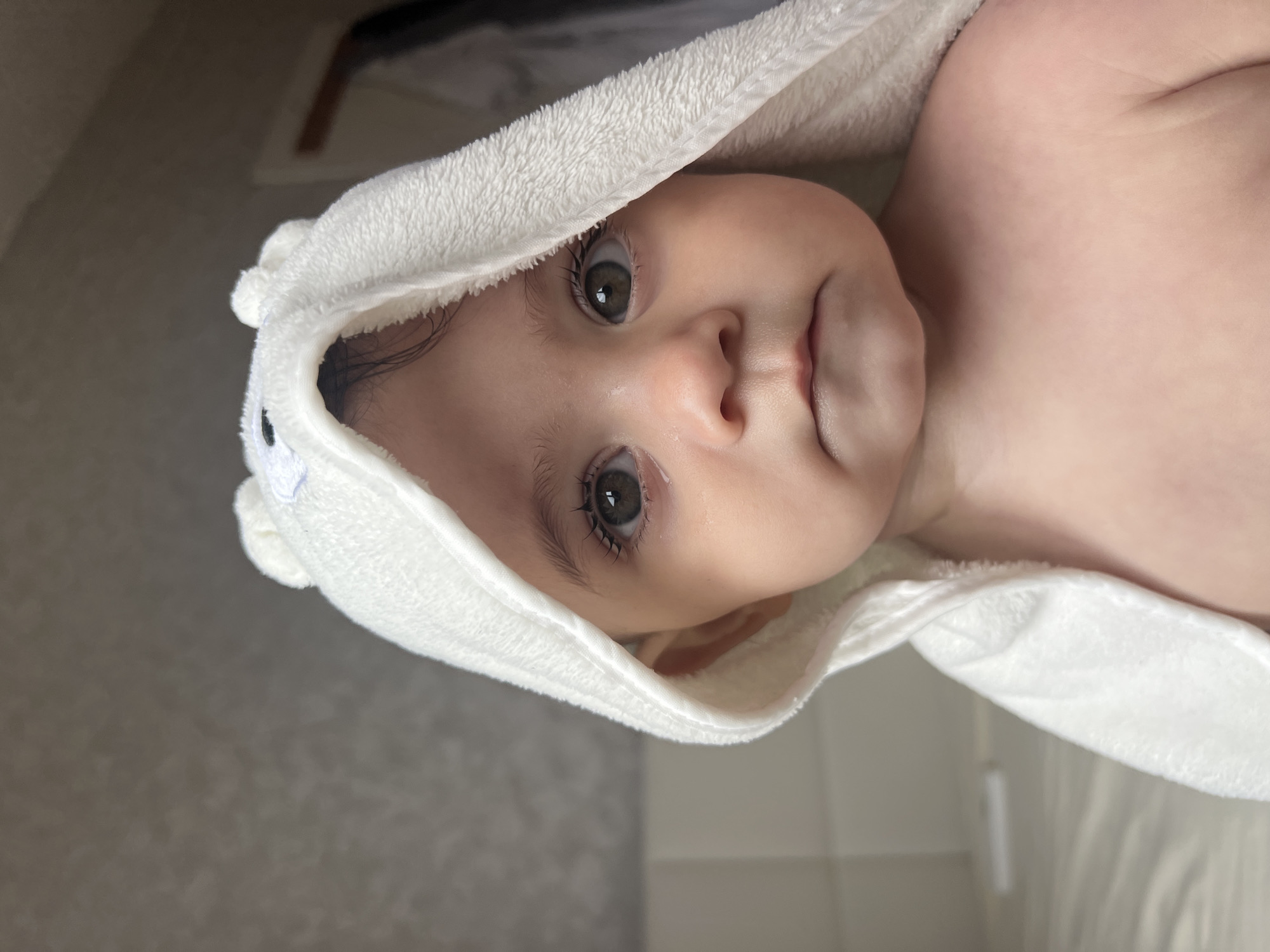 A close-up, medium shot depicts a baby wrapped in a white, hooded towel. The baby's face is the central focus, featuring wide, brown eyes, a small nose, and slightly pursed lips. The towel's hood has small, rounded ears sewn onto it. The baby's bare shoulder is visible on the right side of the frame. The background appears to be a neutral-colored wall, with some possible elements of a bathroom. There is no indication of Elena, Bulgaria.