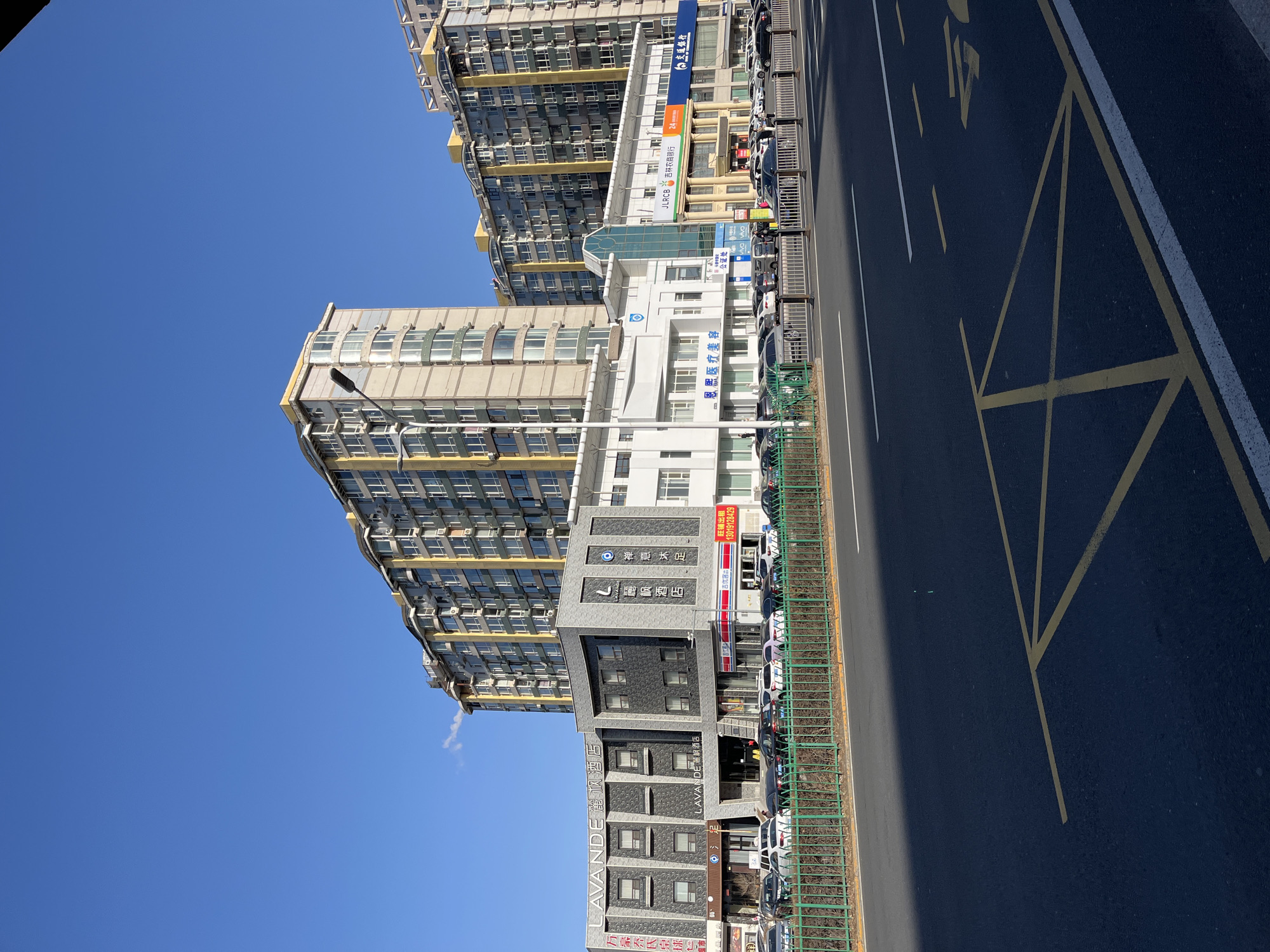 A daytime, medium-shot street-level photograph in Changchun City, China, presents a clear blue sky above several buildings and a road. The tallest building on the left is a high-rise apartment complex with numerous windows, primarily cream and gray with yellow accents. A smaller building to the right, composed of dark gray blocks, displays the name "LAVANDE 麗枫酒店" in Chinese characters.
Another adjacent building is white with a light blue sign that reads "恩熙医疗美容" indicating a medical aesthetic clinic. Further down the street, the building displays the words "JLRCB 吉林农商银行," a Chinese bank. A white traffic line can be seen.
The ground is an asphalt road with yellow painted road markings. A metal green fence runs along the edge of the road. Several cars are parked. The sky is a uniform blue with no clouds, indicating a clear day. A streetlight is visible.