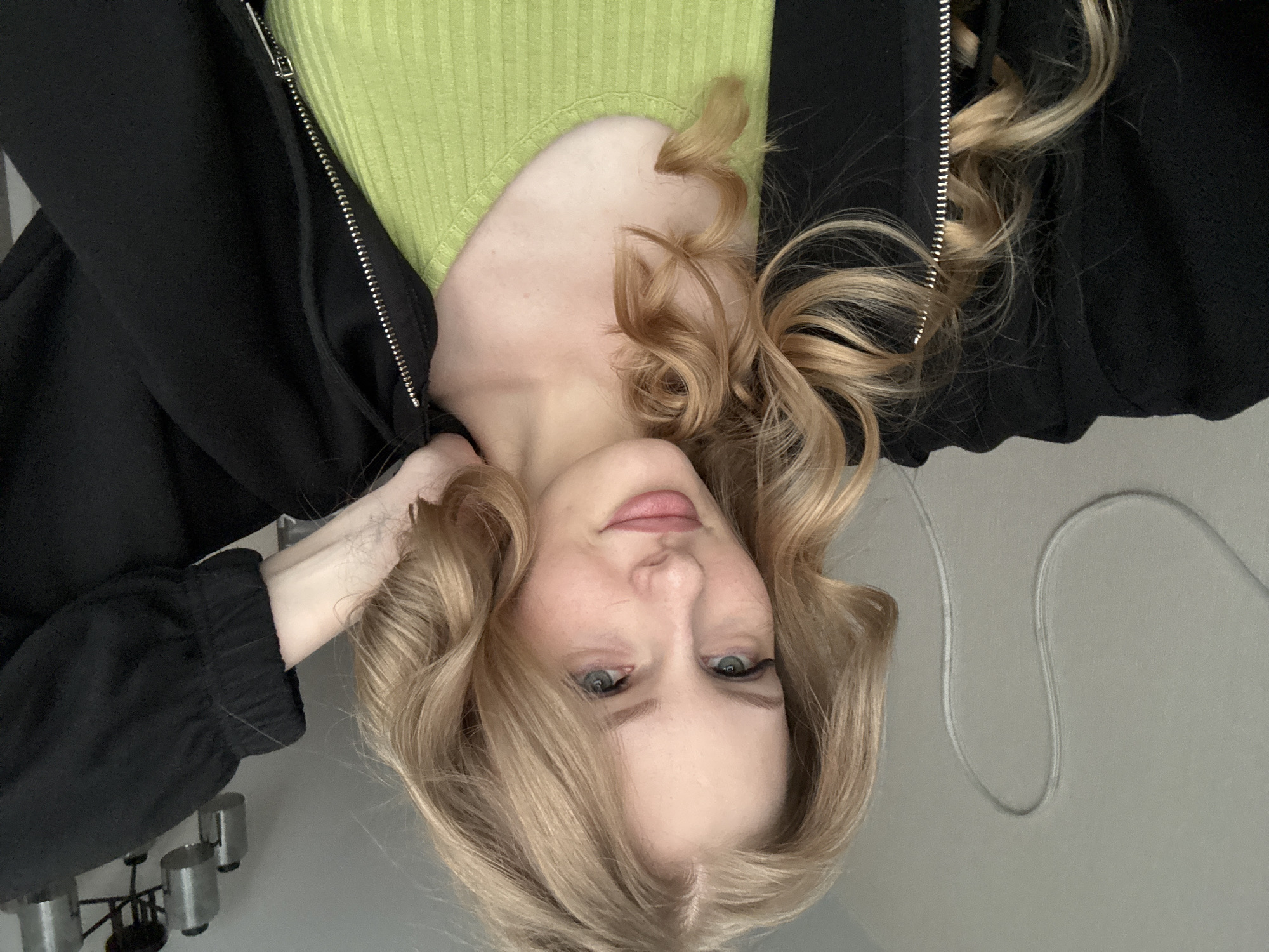 A young woman with blonde wavy hair and light blue eyes is posing for a selfie in an indoor setting. She is wearing a black zippered jacket over a lime green ribbed tank top. Her right arm is raised and her hand is resting on the back of her head. The background appears to be a plain wall with a white cord or wire hanging in a U-shape. A ceiling light fixture with multiple cylindrical lamps is visible in the upper right portion of the image. The overall impression is that of a casual portrait taken in a home environment, possibly in a bedroom or living area. No specific details about the city of Perm, Russia are evident in the image.