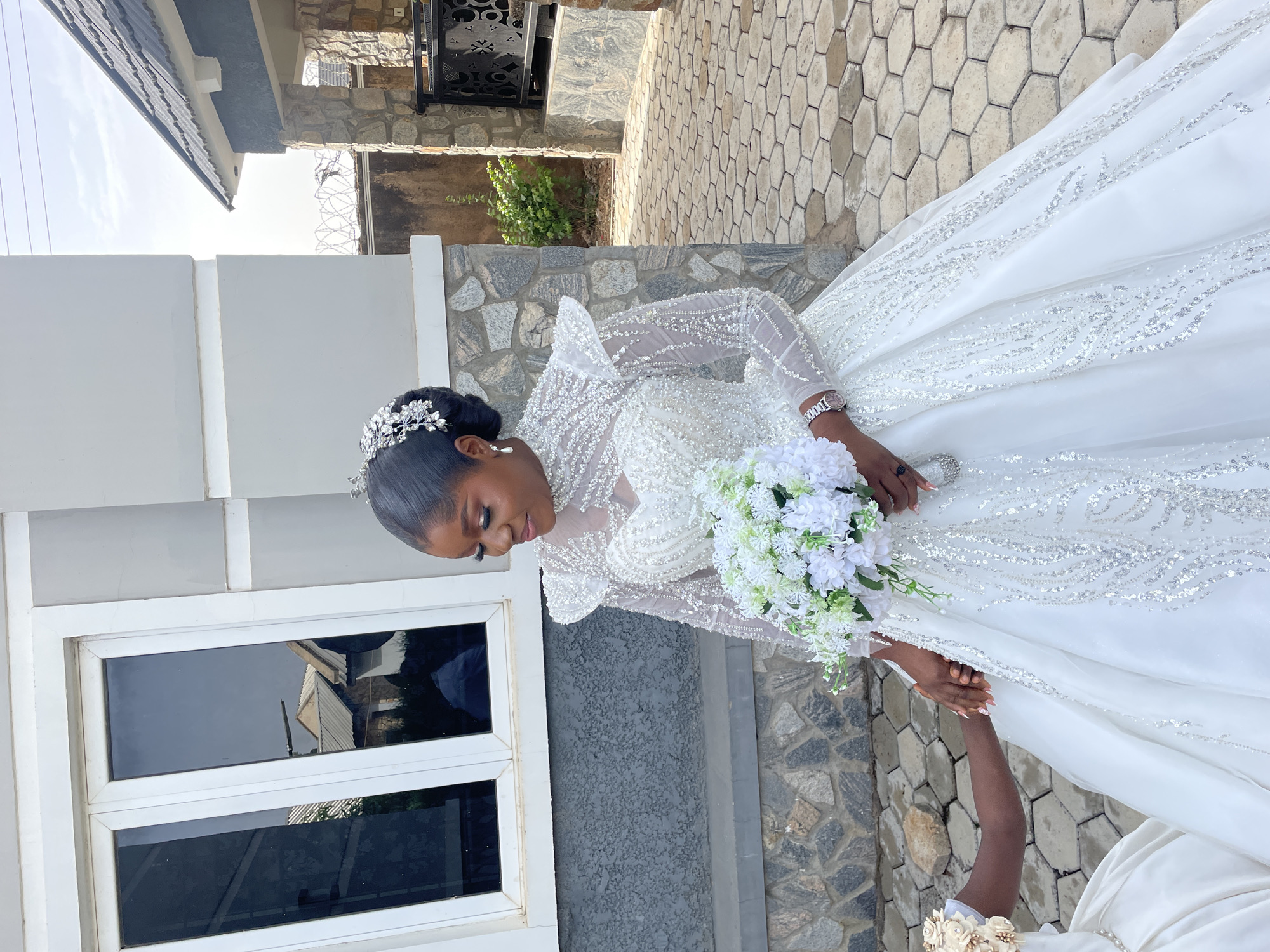 A wedding scene in Bukuru, Nigeria. A woman wearing an ornate white wedding dress with a tiara holds a white floral bouquet, next to a building with stone-textured exterior and white window frames. Another person is holding the wedding dress's edge. The background includes stone pathway, stone walls, and a house.