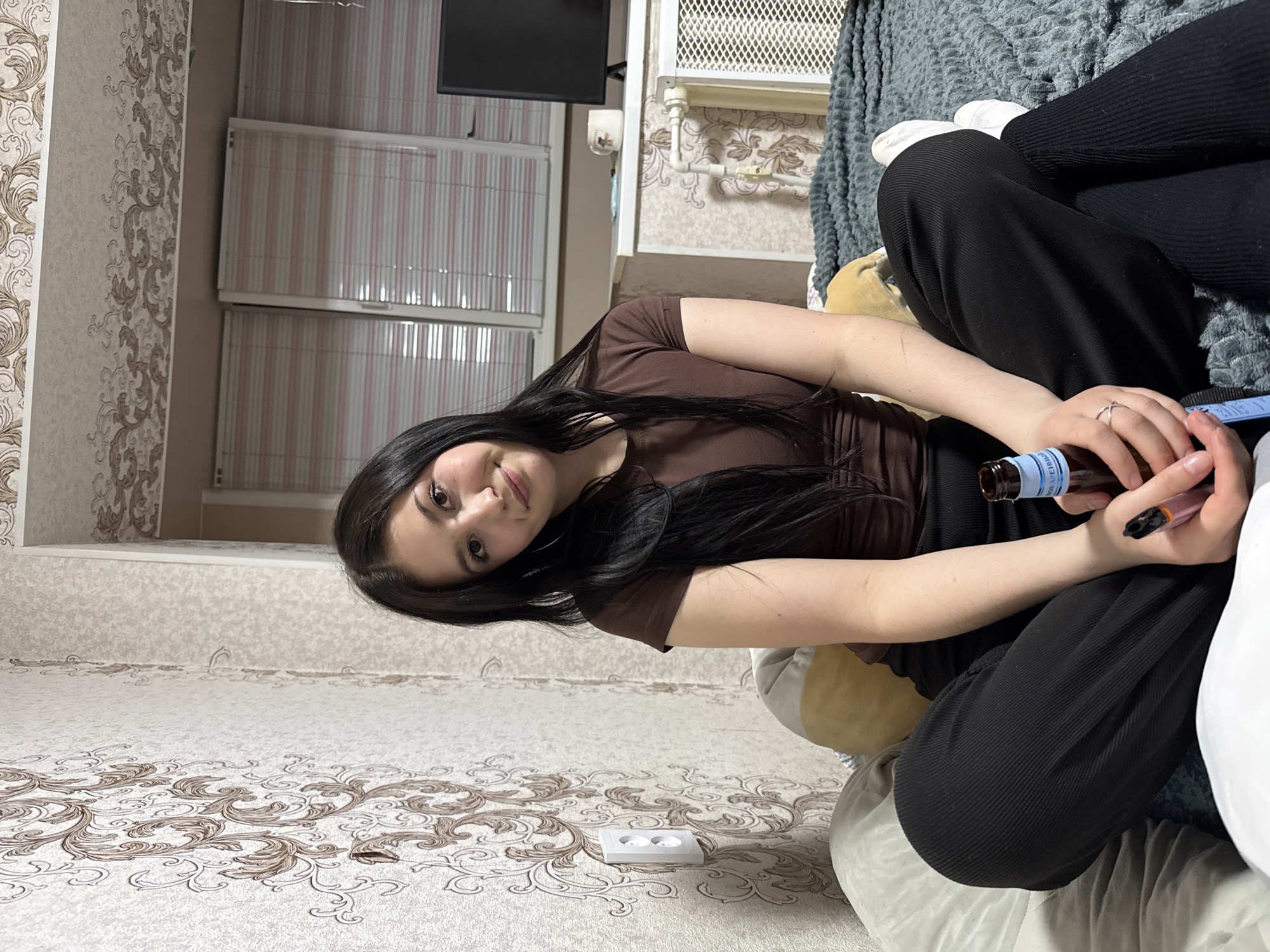 The photograph depicts a young woman with dark hair sitting on a sofa or bed. She is wearing a brown top and black pants. She is holding a bottle and a small device, possibly a vape pen. The background includes patterned wallpaper and radiator covers, suggesting an interior residential space. The photo was taken in Фоки, Russia.