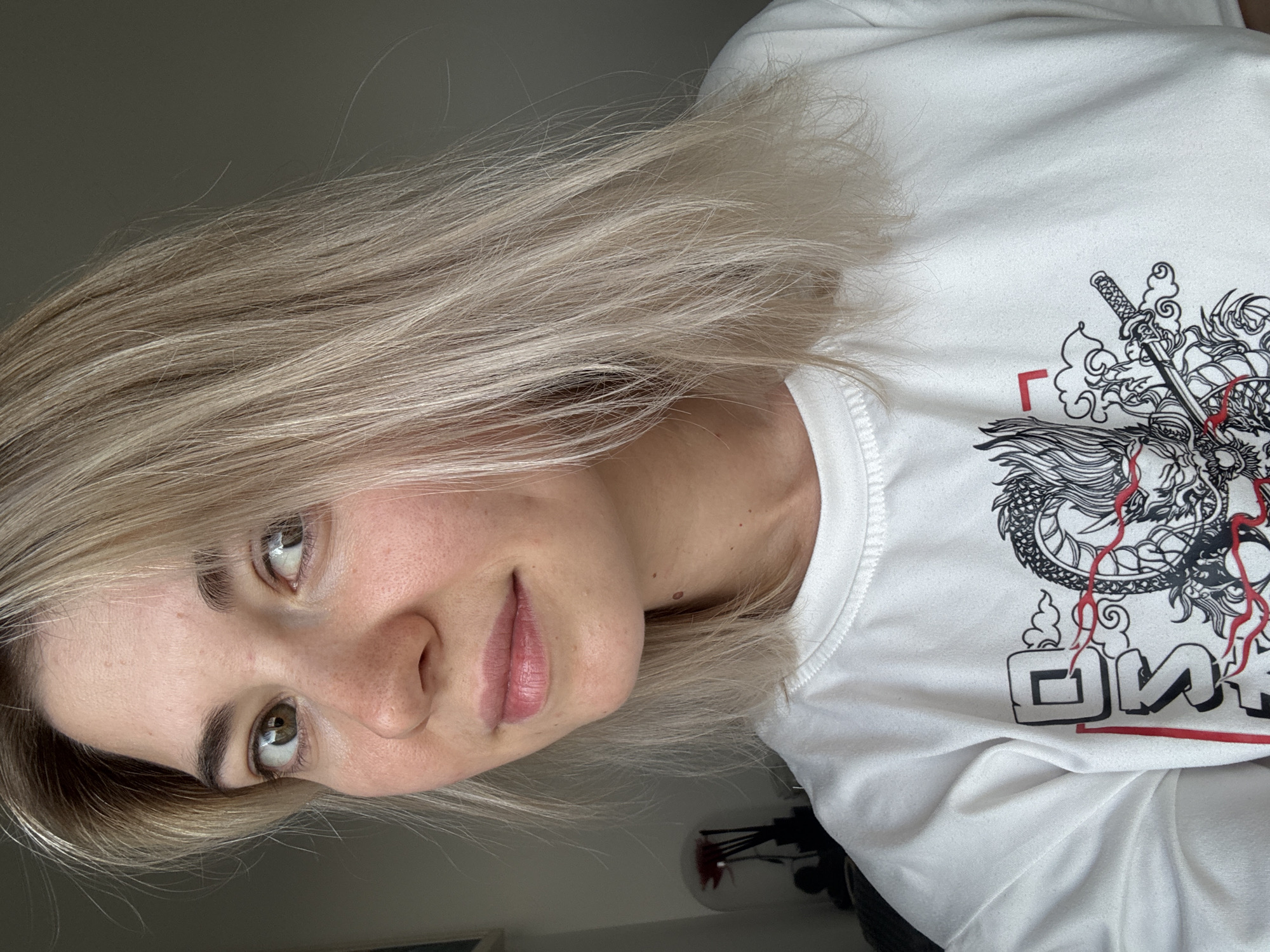 The image shows a close-up, frontal view of a young Caucasian woman with blonde hair and light skin. She is looking directly at the camera with a slight smile. Her eyes are green, and she has light brown eyebrows and a few freckles on her nose and cheeks. She is wearing a white crewneck t-shirt with a black and red dragon design on the front. The background is a plain, dimly lit wall, suggesting an indoor setting, possibly a room in a residence or studio. There is no discernible information about her location in a city.