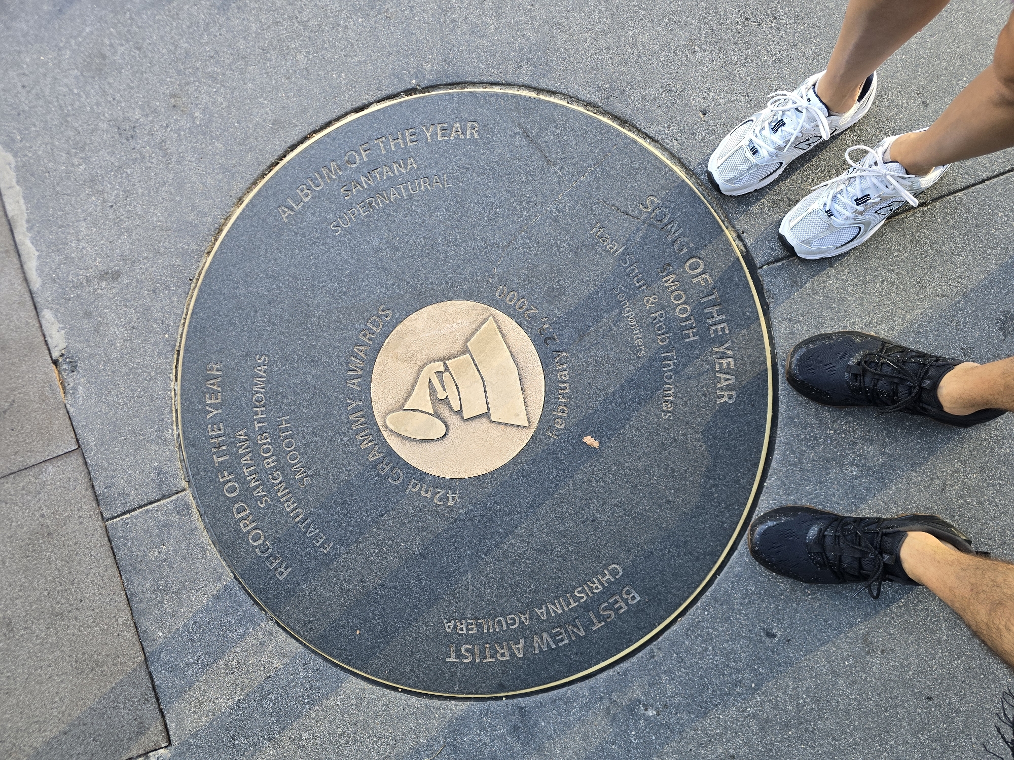 The image shows a close-up, overhead view of a circular bronze plaque embedded in a gray sidewalk. The plaque commemorates the 42nd Annual Grammy Awards and features raised text and a raised image of a Grammy award statuette. Inscriptions around the plaque detail specific awards: "RECORD OF THE YEAR SANTANA FEATURING ROB THOMAS SMOOTH", "ALBUM OF THE YEAR SANTANA SUPERNATURAL", "BEST NEW ARTIST CHRISTINA AGUILERA", and "SONG OF THE YEAR SMOOTH". Below "SONG OF THE YEAR" is text that reads "feat. Stu & Rob Thomas songwriters". The date "FEBRUARY 23, 2000" is also visible.
In the foreground, the feet of two individuals are visible, wearing athletic shoes. One person wears black athletic shoes, and the other wears white athletic shoes. The sidewalk is composed of gray concrete slabs. The scene appears to be outdoors, on a sunny day, and the plaque is located in Los Angeles, California, USA, as it is part of the Hollywood Walk of Fame, specifically near the Dolby Theatre where the Grammy Awards were held annually at that time.