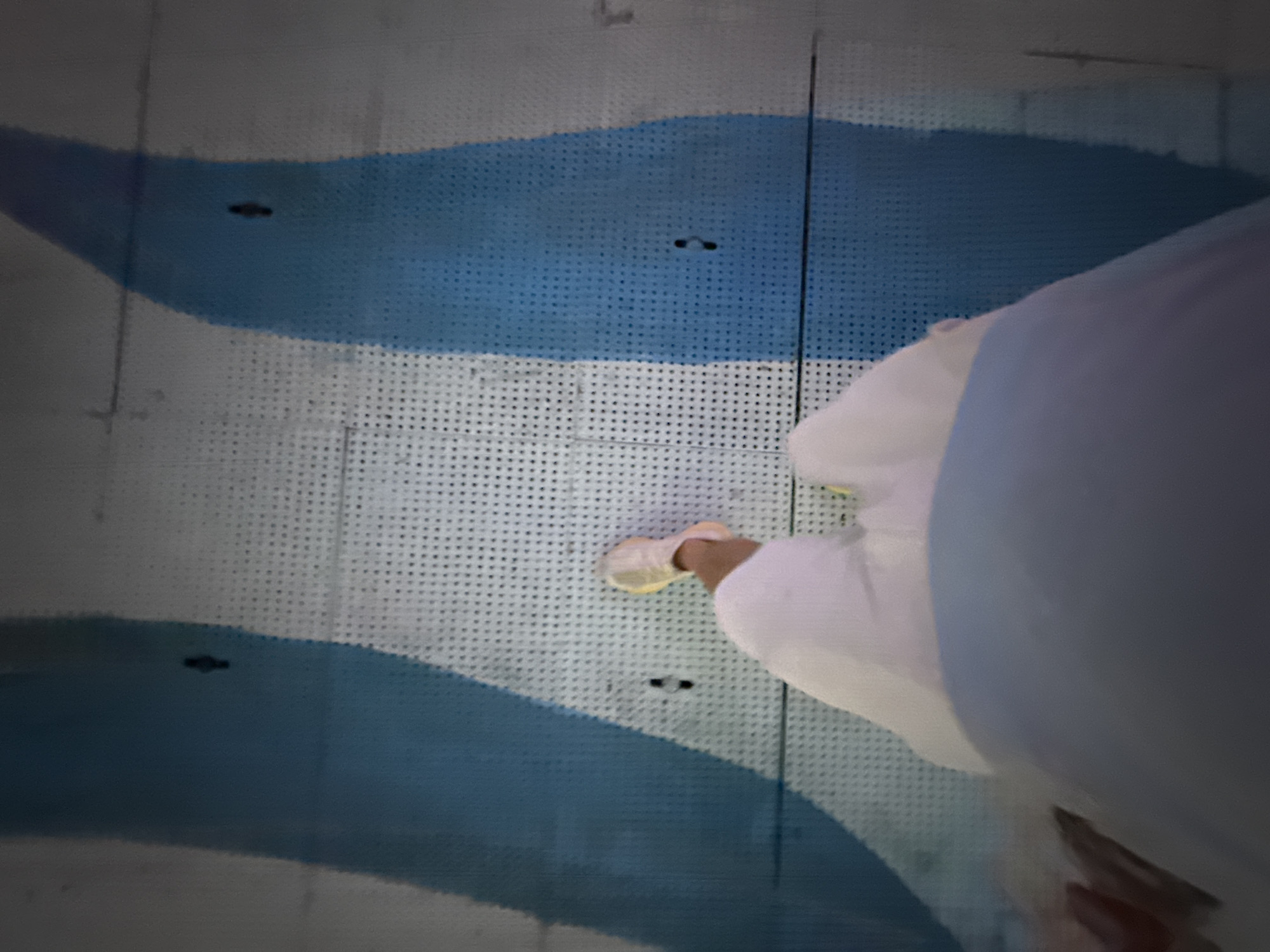 A person wearing white clothing and light-colored sneakers walks on a surface with a perforated grid pattern. The surface is decorated with two wide, wavy bands of blue. The perspective is from above, looking down at the person's feet and lower legs. The blue bands appear to be part of a mural or painted design on the ground or a wall. The lighting suggests it might be indoors or a shaded outdoor area, as there are no strong shadows indicative of direct sunlight. The overall scene is abstract due to the focus on the pattern and the person's attire. There is no visible text in the image. The location context is Tianhe District, China, but there are no specific visual cues within the image to confirm this.