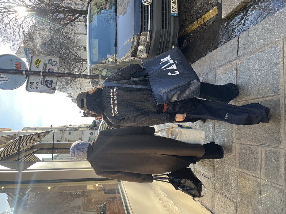 Three individuals are depicted walking away from the camera on a stone-paved sidewalk in a daytime urban environment. The location is identified as Paris, France, based on the architecture, street signs, and a French vehicle license plate visible.
The leftmost individual wears a dark brown long coat over dark trousers and a light-colored striped beanie. They carry a black shiny bag in their left hand and a white tote bag with colorful graphics in their right hand.
The middle individual is partially obscured, wearing dark clothing.
The rightmost individual wears a dark quilted bomber jacket with white text on the back: "Automne-Hiver '84-'85 COMME des GARÇONS." They wear dark, loose-fitting trousers, with "Helly Hansen" branding visible on the left leg, and a red belt. A brown baseball cap is worn backward over dark, mid-length hair. This person carries a large dark blue tote bag with "CALM" printed in white, suspended from their right shoulder by a woven strap.
To the right of the pedestrians, a dark Volkswagen van is parked parallel to the curb on an asphalt street. A metal pole with two circular street signs, obscured by multiple stickers including "SOSA" and "REMY DAWG," stands between the sidewalk and the street. The street surface includes a yellow painted line near the curb, where some liquid is visible in the gutter. Buildings featuring classic multi-story Parisian architecture line the street; the building immediately to the left of the pedestrians has large display windows showcasing merchandise and a striped awning. Bare tree branches are visible against a bright sky in the background.
