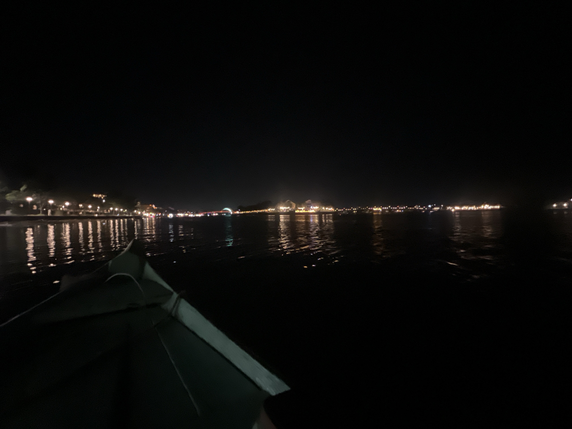 An outdoor, wide shot taken from a boat at night shows a dark body of water with a distant city skyline illuminated by numerous lights. The boat, partially visible in the foreground, is dark green and appears to be made of fiberglass. The city lights are reflected in the water, creating streaks of light. The scene is dark and quiet, with the exception of the distant city lights. The location is likely the Thu Bon River in Hoi An, Vietnam, known for its nighttime boat tours and illuminated ancient town.