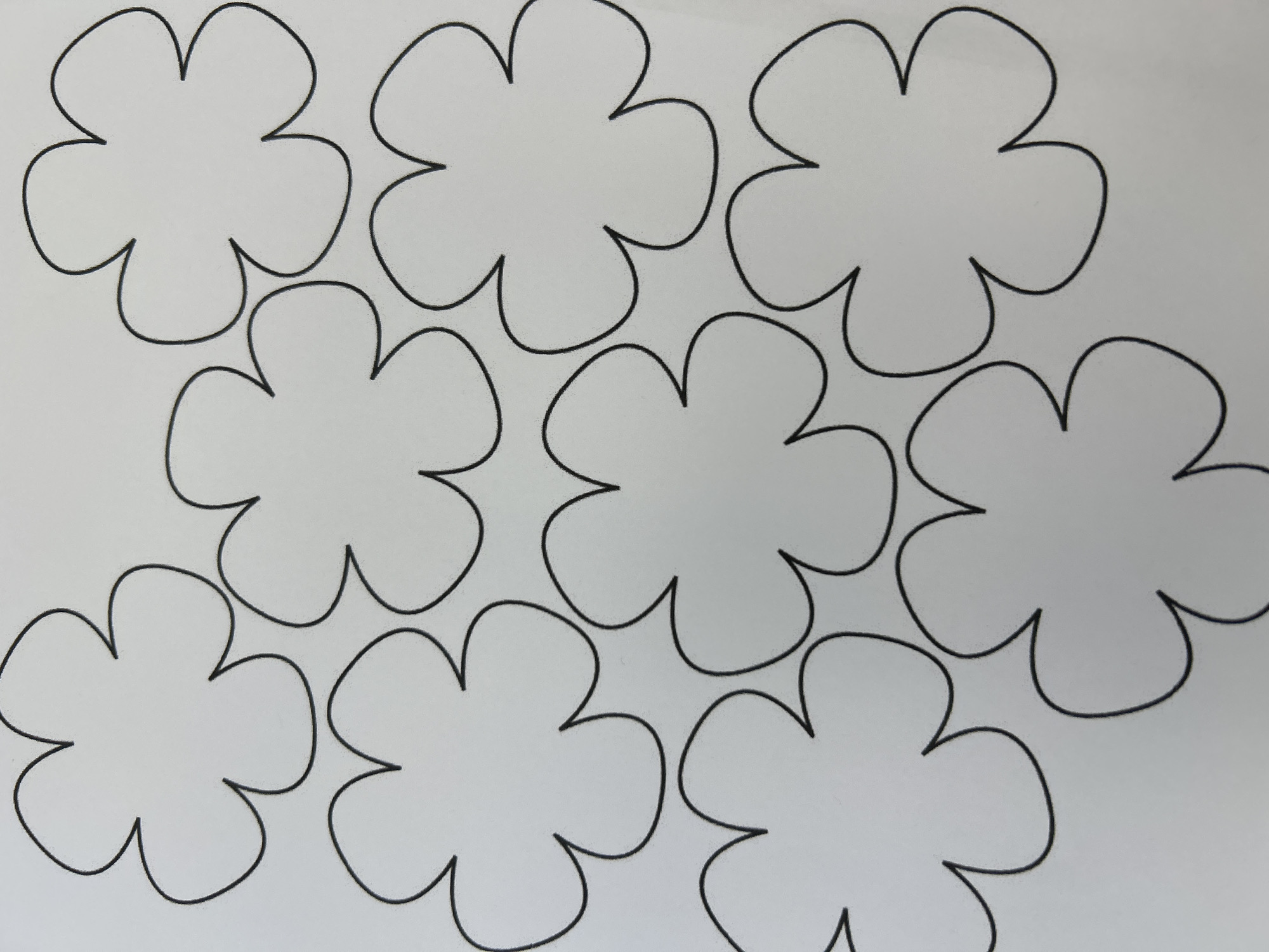 A white sheet of paper displays an arrangement of nine black outlines of flowers. The flowers are abstract, each possessing five rounded petals radiating from a central point. They are scattered across the page in a somewhat disorganized but visually balanced pattern. The lighting is even, and the image is taken from a top-down perspective, indicating it is likely a photograph of a drawing or stencil. There are no people, city elements, or environmental details present beyond the paper itself.