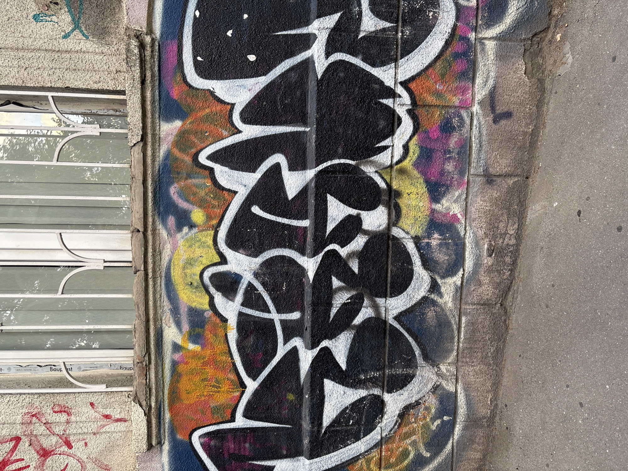 The image displays a section of an urban wall covered in vibrant graffiti. The graffiti features bold, black lettering outlined in white, creating a dynamic and eye-catching design. Behind the main lettering, splashes of colorful spray paint in orange, yellow, pink, and blue add depth and texture to the artwork. The wall itself appears to be made of stone or brick, with visible grout lines. To the left of the graffiti, a window with horizontal metal bars and reflective blinds is visible, suggesting an indoor space is behind it. The lower portion of the wall shows some red graffiti tags. The ground to the right of the wall is asphalt. The lighting suggests it is daytime, with sunlight casting shadows. The scene depicts a moment of urban art and decay, with the graffiti overlaying the textured surface of the building. There is no clear indication of people or a specific event taking place. Some faint text, "Baup" and "st", is visible on the window frame.
