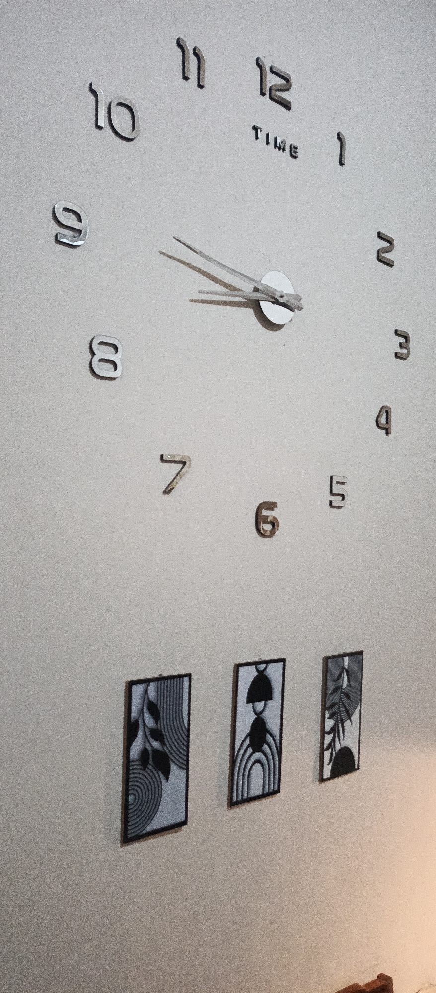 The image depicts a wall-mounted clock with three framed pictures below it. The clock has silver numbers for hours, with the number 12 having the word "TIME" written beneath it. The clock hands are silver and are positioned between the 10 and 11. The three framed pictures are vertically oriented and feature abstract black and white designs. The picture on the left has a pattern of curved lines and leaf-like shapes. The middle picture displays a half-circle shape above a series of concentric arcs. The picture on the right shows a branching leaf motif. The wall is a light, neutral color. No people are visible in the image. The scene appears to be an indoor setting.