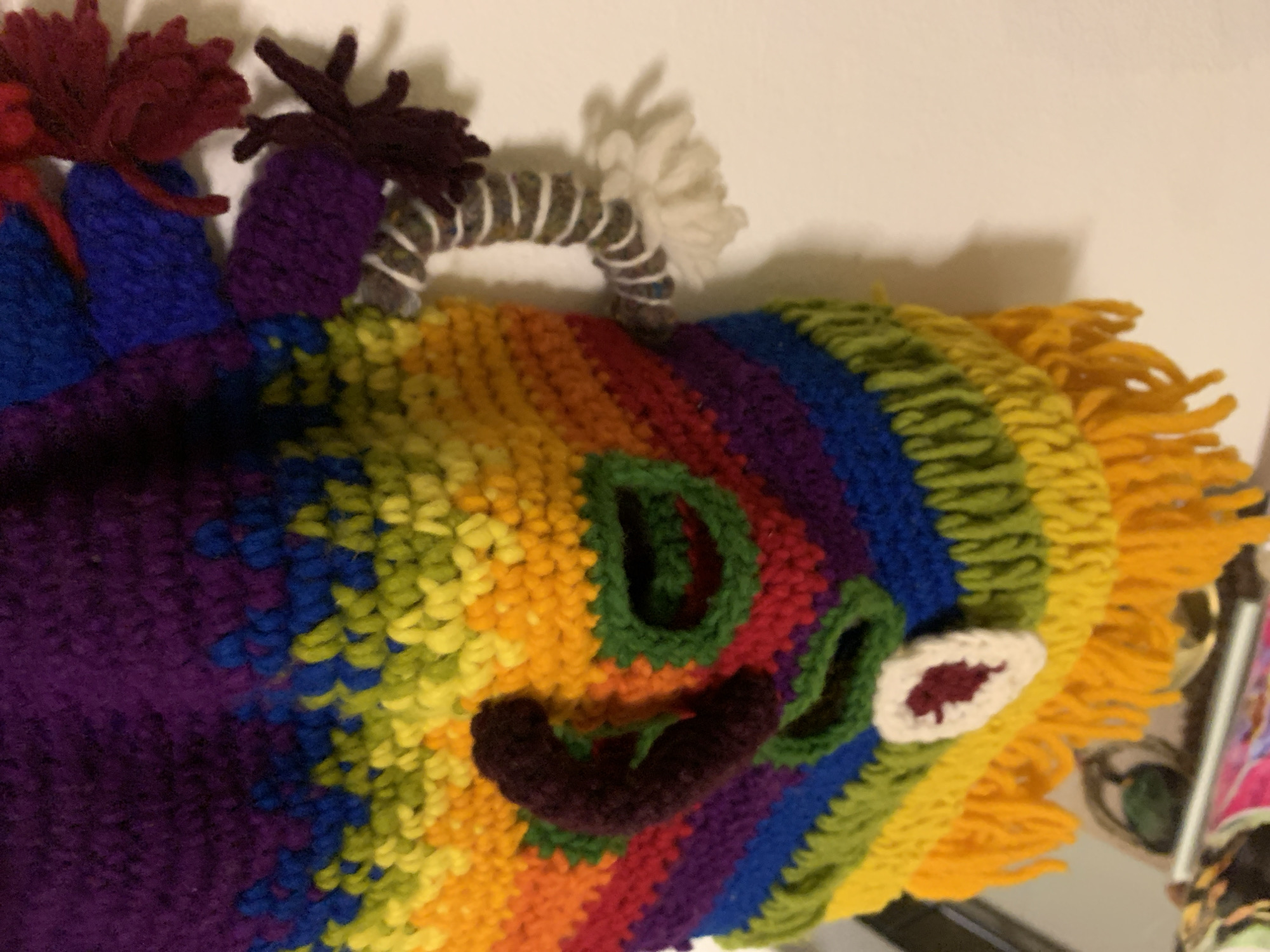 An overhead close-up shot shows a colorful, crocheted mask with various facial features. The mask is made of yarn and features stripes of purple, blue, yellow, orange, red, and green. There is a dark brown, curved nose-like appendage on the left side of the mask, and green crocheted lips. A white oval shape with a dark red center is on the lower right of the mask, possibly representing an eye or cheek marking. On the right side of the mask, a coiled, arm-like appendage made of alternating brown and white yarn is attached. This appendage ends in a fluffy white pom-pom. The mask is hanging against a plain, light-colored wall. The bottom of the mask is fringed with yellow yarn.