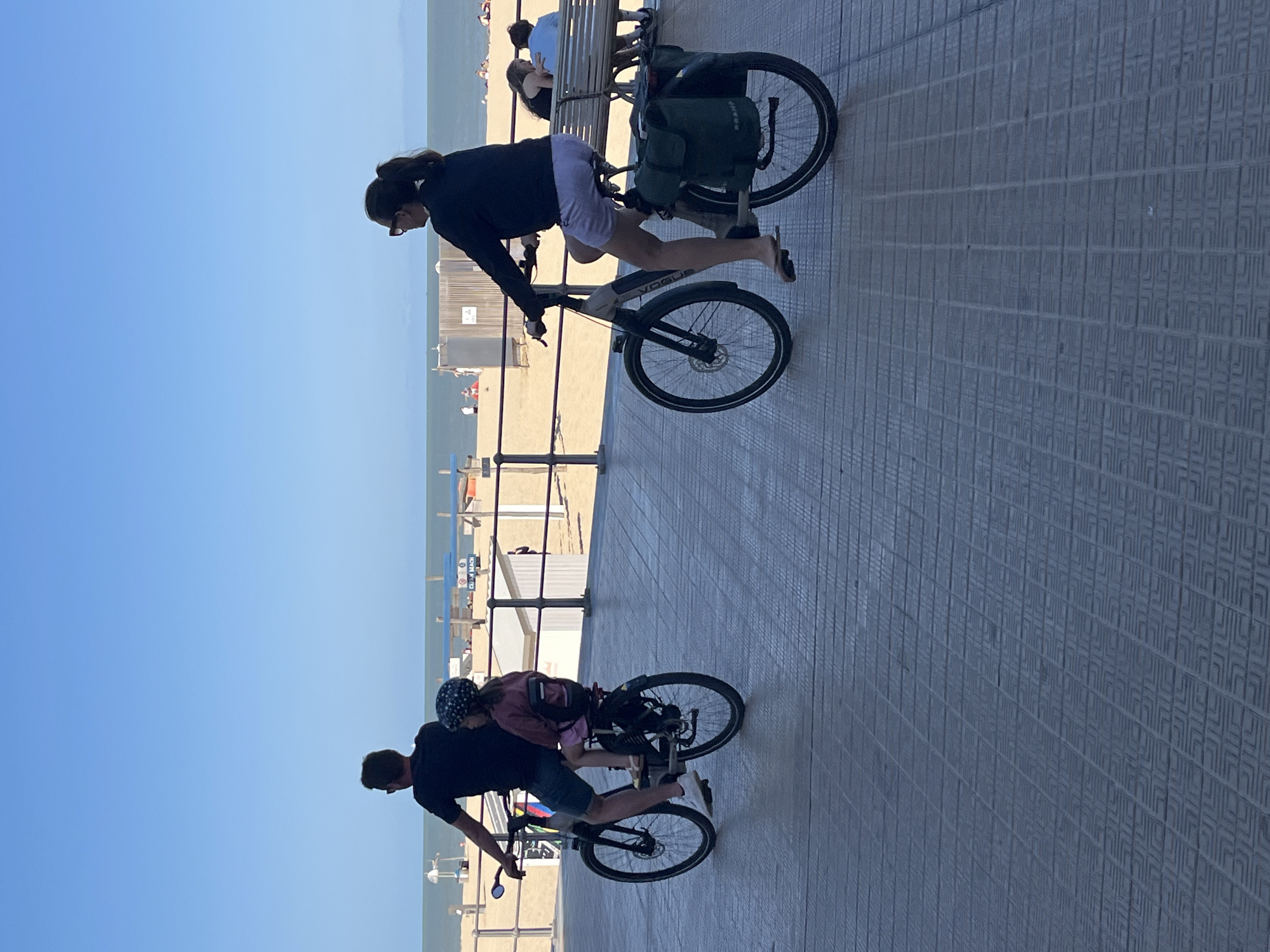 In Ostend, Belgium, on a sunny day, a man and a child ride a bicycle together, while a woman rides another bicycle beside them on a paved walkway. The walkway borders a sandy beach with a blue sky overhead. The ocean is visible in the background, with a few people scattered on the beach and in the water. The man wears a dark t-shirt and shorts, while the child is in a pink shirt and a helmet. The woman is in a black top and light purple shorts, with a green bag attached to her bicycle. The scene depicts a leisurely outing at the beach.