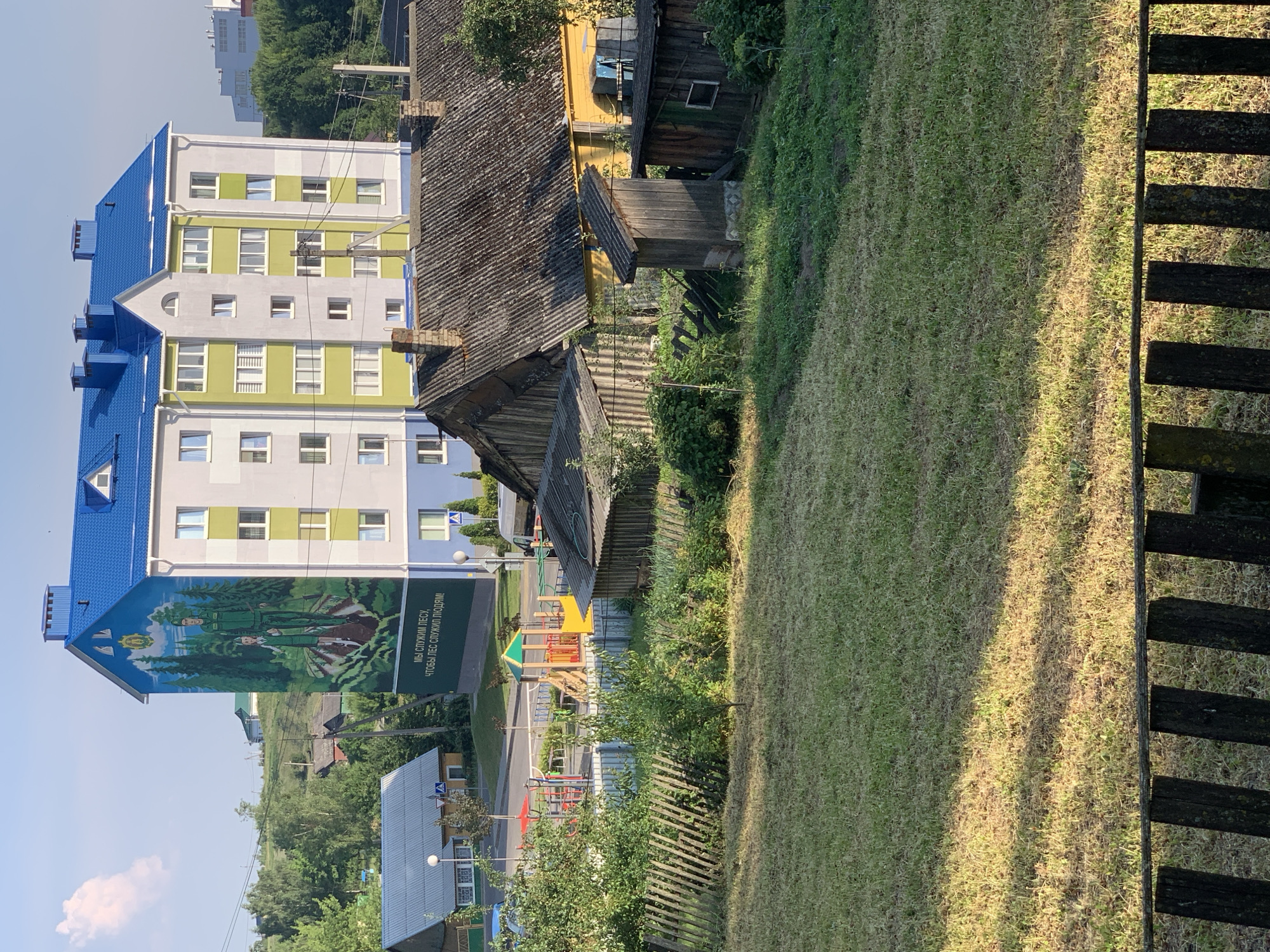 This media file shows an exterior view of a multi-story building in Kapyĺ, Belarus. The building features a striking mural on its side, depicting two figures in military uniforms surrounded by trees. The text on the mural reads "МЫ СЛУЖИМ ЛЕСУ, ЧТОБЫ ЛЕС СЛУЖИЛ ЛЮДЯМ!" which translates to "WE SERVE THE FOREST SO THAT THE FOREST SERVES PEOPLE!".
To the left of the building, a modern apartment complex with a bright blue roof and green and white facades is visible. In front of this complex, there is a children's playground with colorful equipment. Adjacent to the apartment complex, older, rustic wooden structures with corrugated roofs stand, suggesting a mix of old and new architecture in the area.
The scene is bathed in the warm light of what appears to be either early morning or late afternoon, indicated by the long shadows cast across the grassy slope and wooden fence in the foreground. The weather seems to be fair, with a clear sky. The overall atmosphere suggests a peaceful, everyday scene in a residential area.