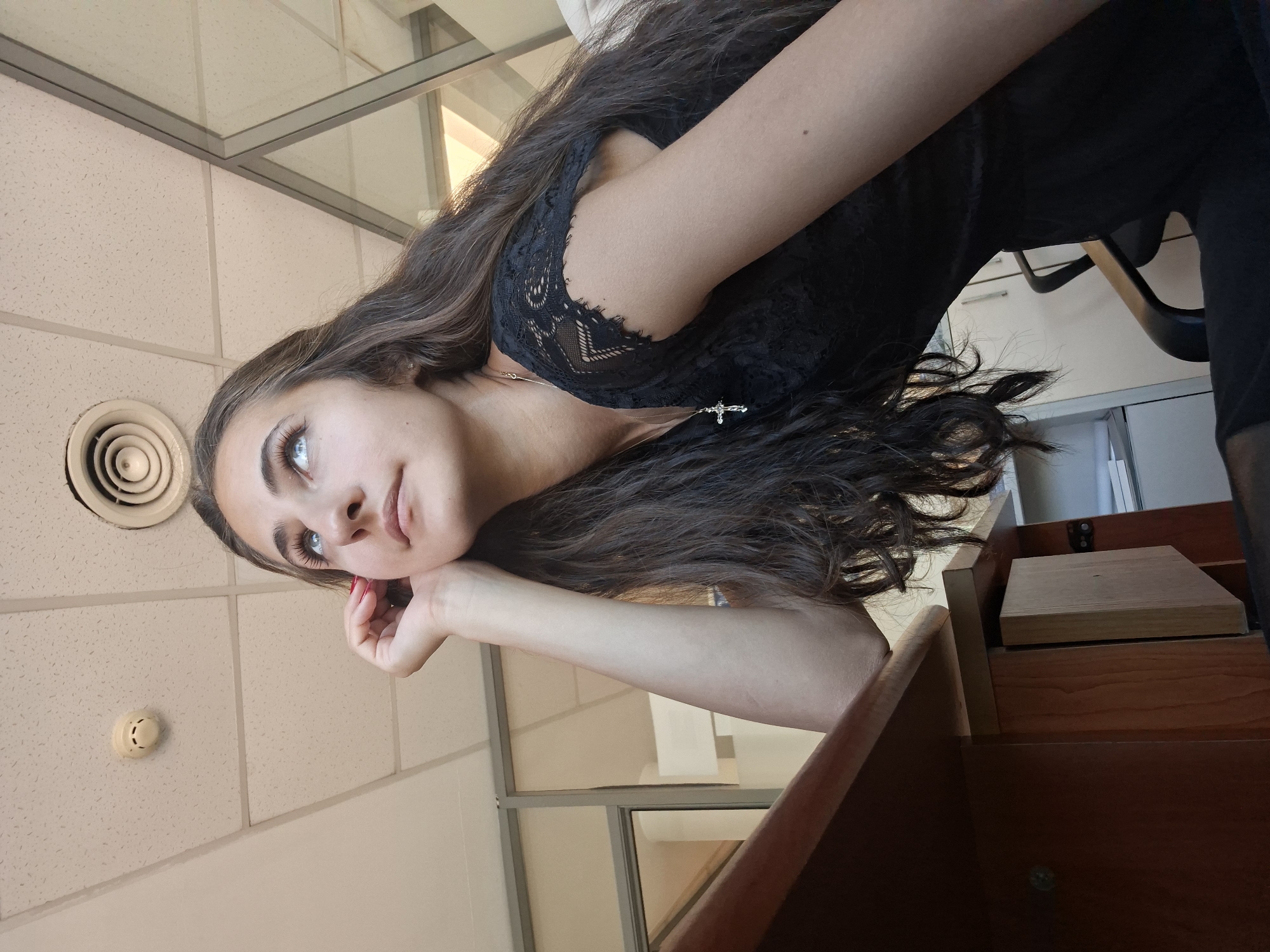 A young woman with long, dark, wavy hair is pictured in a medium shot, leaning her head on her left hand and looking upwards. She is wearing a black lace top and a necklace with a cross pendant. The background shows an office environment with a drop ceiling, a smoke detector, an air vent, a wooden desk, and office chairs and cabinets. The lighting suggests it is daytime. The location is an office space, though the specific city and country are unknown.
