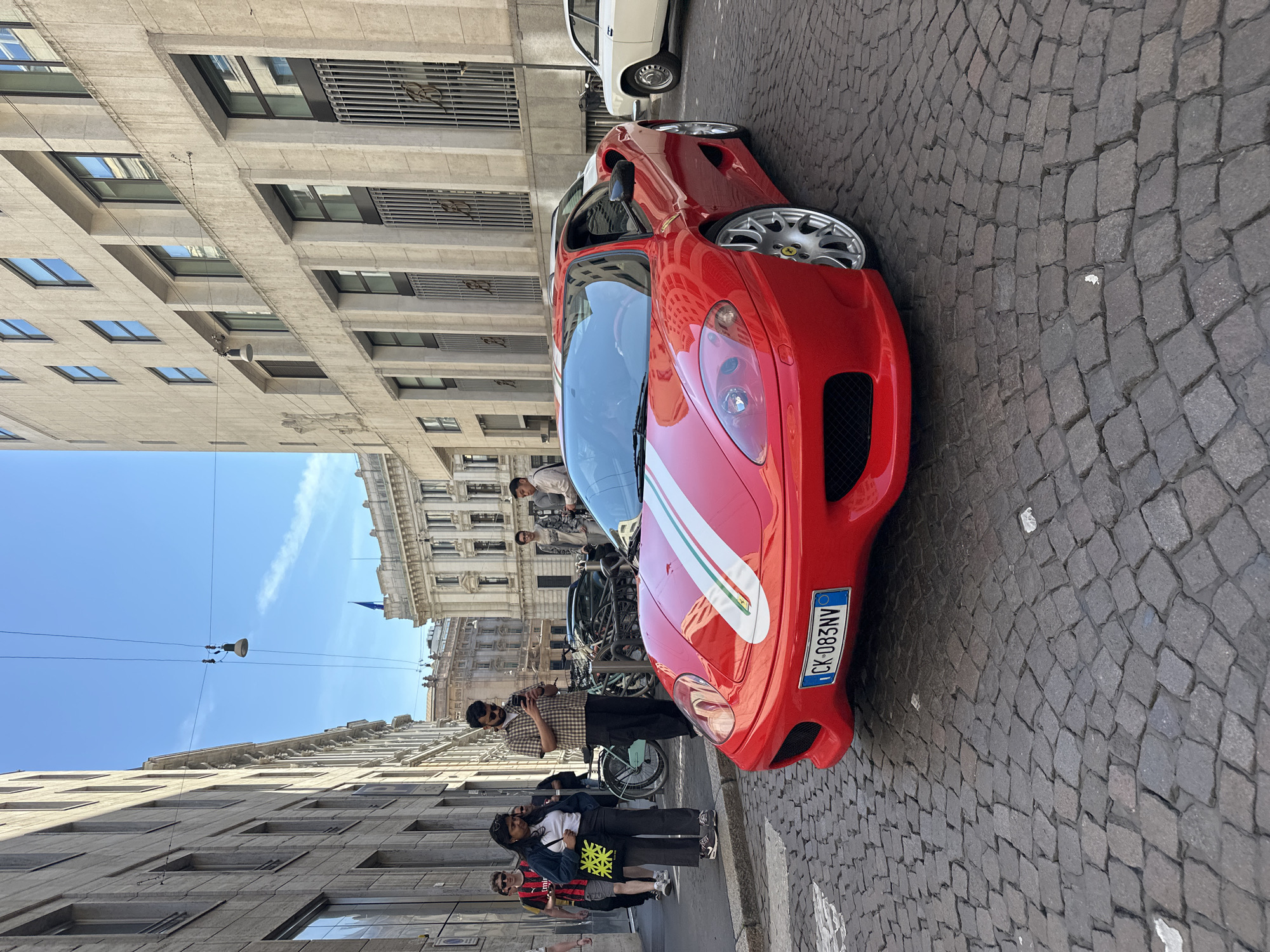 The image depicts a red Ferrari 360 Modena parked on a cobblestone street in Milan, Italy. A white stripe with Italian flag colors runs along the center of the hood. A partially visible license plate reads "CK-083NV". Several people are standing near the car, including a man holding a phone, potentially taking a picture of the Ferrari. Buildings with multiple windows line the street, reflecting the sunlight. Overhead, streetlights hang from wires between the buildings. Other parked vehicles are visible further down the street.