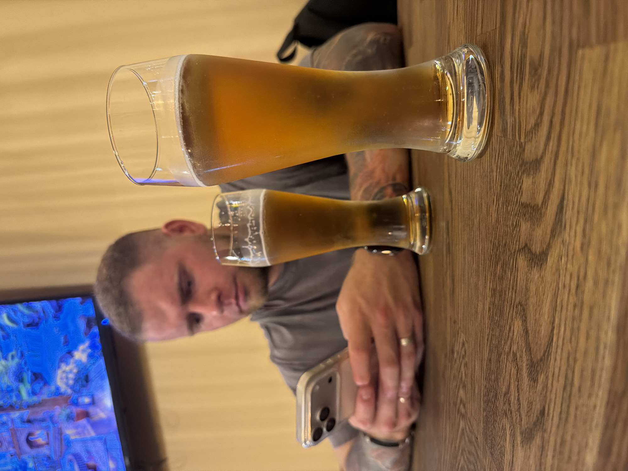 A man with short hair and tattoos on his arms is sitting at a wooden table, looking at his phone. He is wearing a gray t-shirt. In front of him are two glasses of beer. The background is a light yellow wall, with a television screen displaying a colorful image in the upper left corner. The scene appears to be indoors, possibly at a restaurant or bar, though no specific Chaikovsky landmarks are visible. The lighting is dim, with a warm glow.