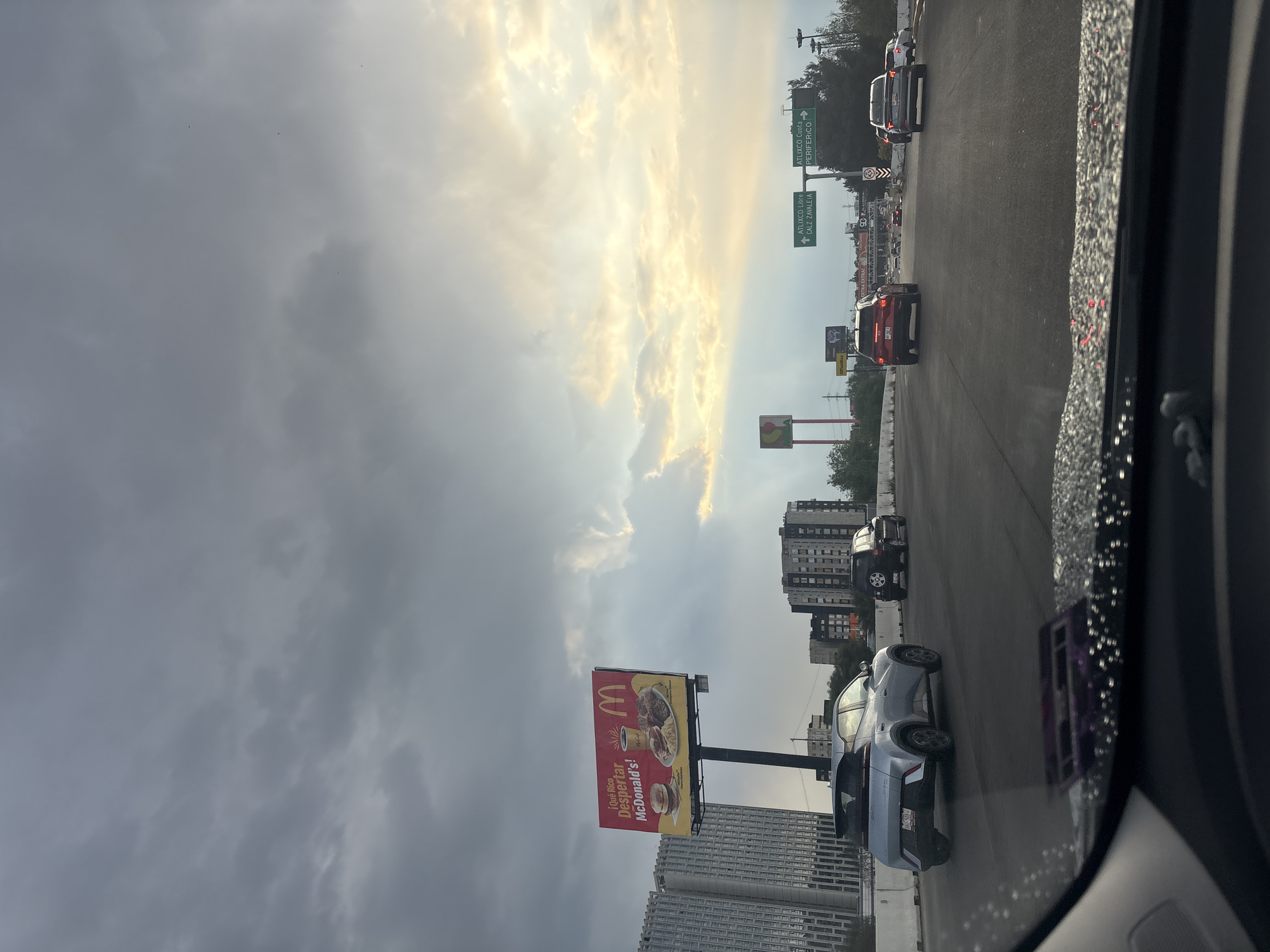 The image, taken from inside a vehicle in San Bernardino Tlaxcalancingo, Mexico, shows a highway during daytime under a cloudy sky with a visible patch of bright sunlight. Cars are traveling in the lanes adjacent to the one the vehicle is in. A McDonald's billboard is positioned along the roadside, featuring Spanish text. Road signs with green backgrounds, pointing towards "ATLIXCO Libre" and "ATLIXCO Cuota PERIFERICO," are visible in the distance. Buildings are seen along the highway. The windshield has water droplets on the right side, suggesting recent rain or cleaning.