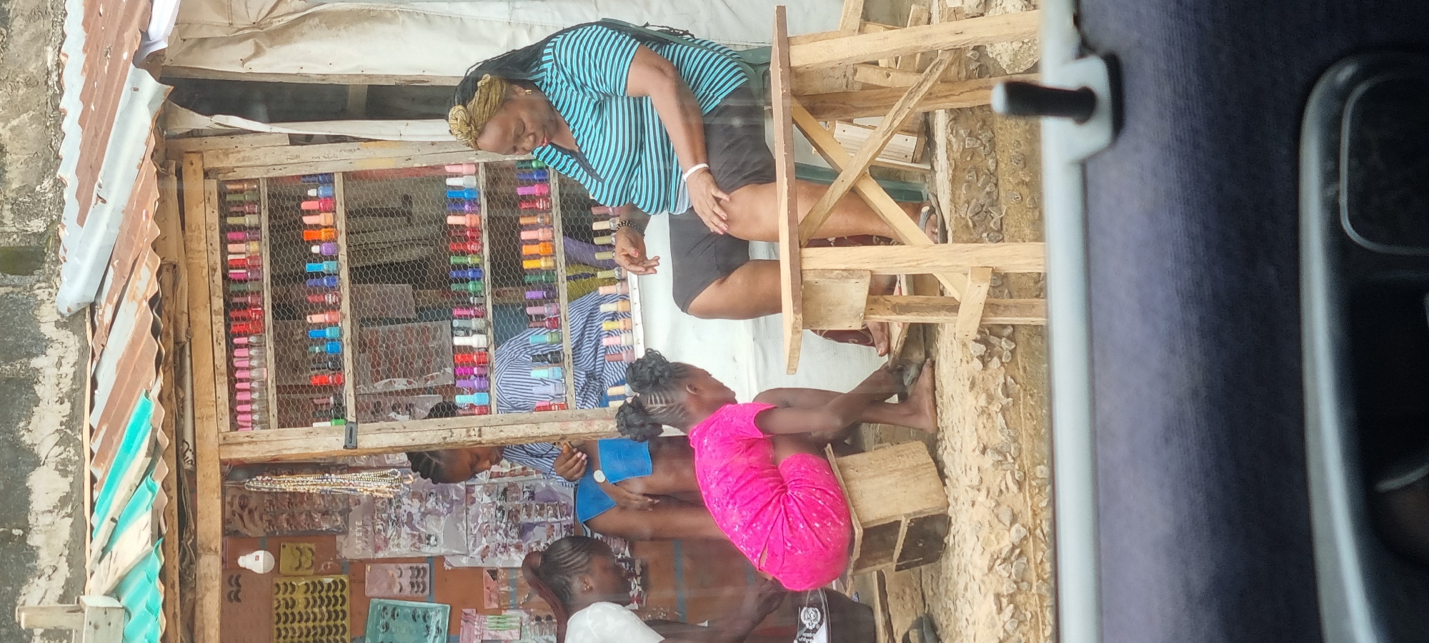 In Lagos, Nigeria, a street vendor operates a nail salon from a makeshift stall. The stall features shelves stocked with an array of colorful nail polish bottles. A woman with braided hair, dressed in a blue and white striped shirt, is seen working on a customer's nails. Another woman, wearing a bright pink dress, sits on a wooden stool and appears to be having her nails done as well. A larger woman with a blue and green striped shirt and a headwrap sits on a wooden chair, observing the activity. The scene is outdoors, with a rough, gravelly ground and a corrugated metal roof covering the stall, indicating a typical street-side business in Lagos.