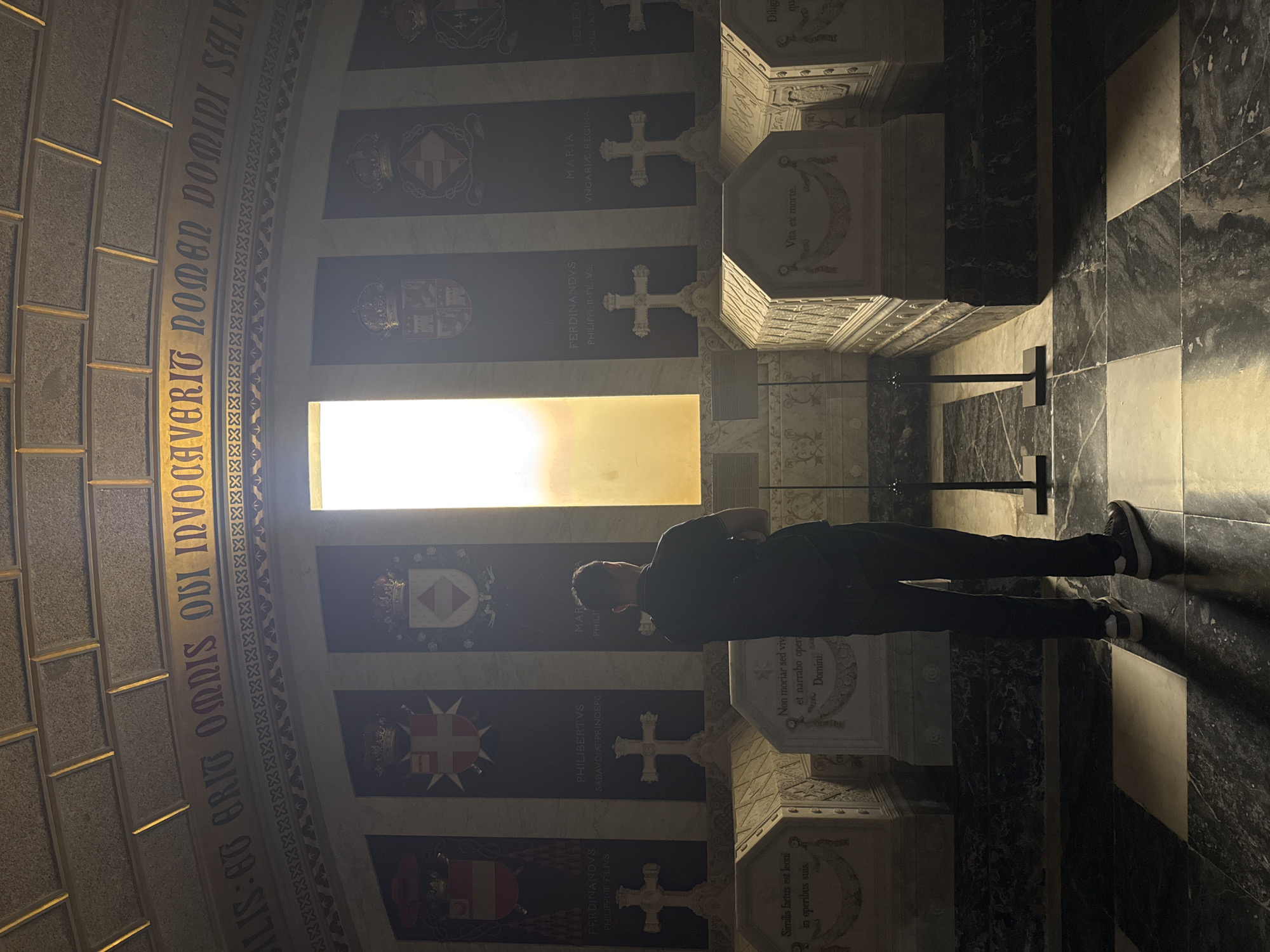 A man stands in the Royal Pantheon of San Lorenzo de El Escorial, Spain, facing a tall, narrow window that emits bright, diffused light. The man, silhouetted against the light, is viewed from behind, wearing a dark t-shirt and dark pants, with white-soled athletic shoes. He has a backpack on. The Pantheon is dimly lit, with the man's presence being the primary human element.
The space features a vaulted ceiling with visible rectangular panels. A decorative golden inscription runs along the upper wall, with the visible text reading "IS: ET GRIT OMNIS QUI INVOCAVERIT NOMEN DOMINI SALV" and continuing further along the curve. Below this, a decorative border of interlocking X shapes is present.
Flanking the window are several dark rectangular panels, each adorned with a coat of arms topped by a crown. Below these are large white crosses. These are set above rows of sarcophagi, which are ornate and carved, with inscriptions. One sarcophagus visible on the left has an inscription that appears to read "Similis factus est leoni in operibus suis." Another in the center reads "Non moriar sed vivam et narrabo opera Domini." A third on the right has the inscription "Vita ex morte." The sarcophagi are made of white marble and are placed on a dark, polished marble floor with a checkerboard pattern of light and dark squares. The walls behind the sarcophagi are also of dark, polished marble. Two tall, thin black poles with square bases are positioned in front of the sarcophagi, likely acting as barriers or for display.