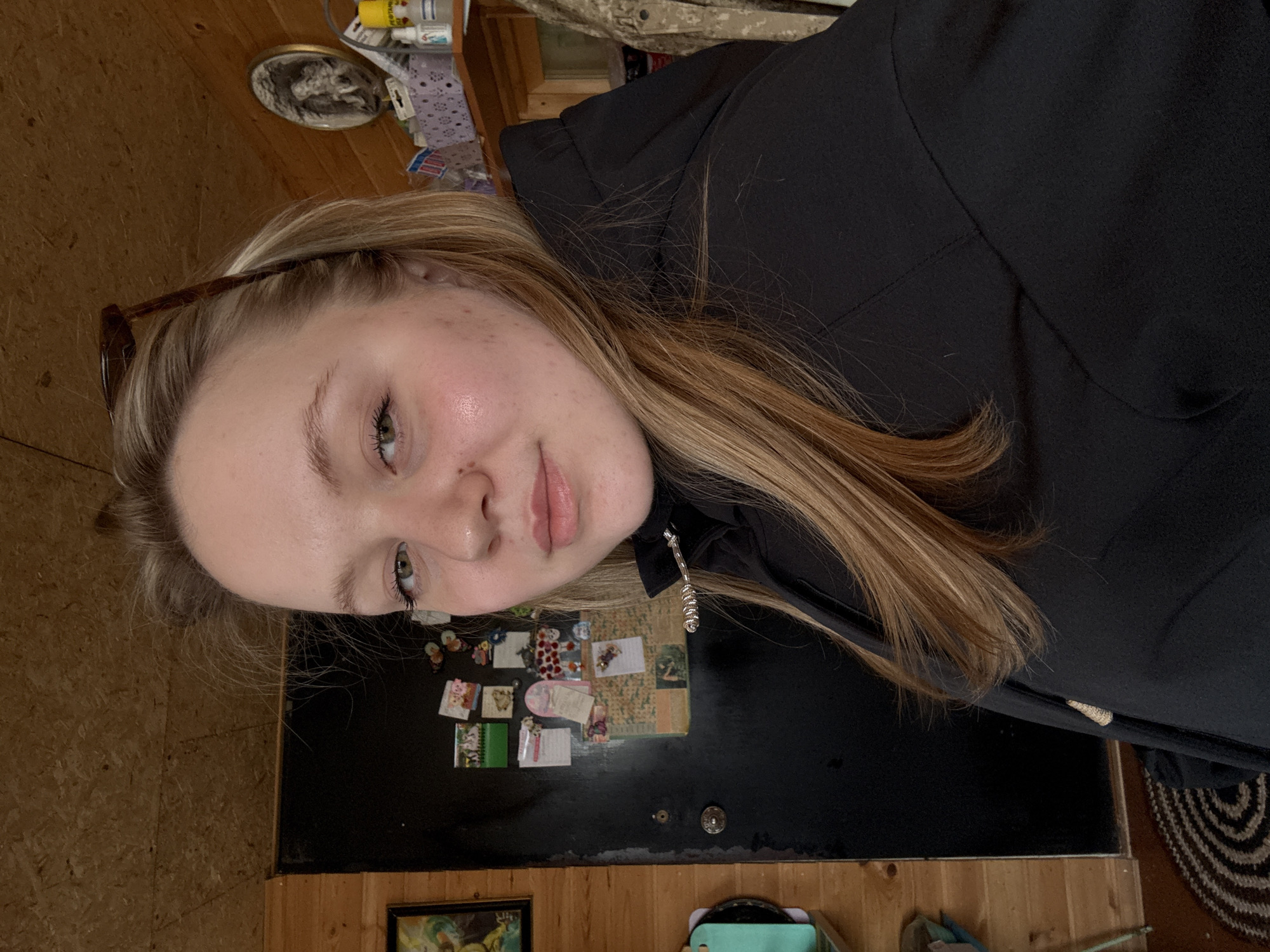 A young woman with blond hair is the subject of the photograph. She has light skin and is wearing a black hooded sweatshirt. The setting appears to be indoors, with a wood-paneled wall visible. A black surface with various items attached, including small notes, stickers, and images, is also in view. The location is Гаревая, Russia, but the image itself does not offer any specifics to pinpoint the exact area within the city.