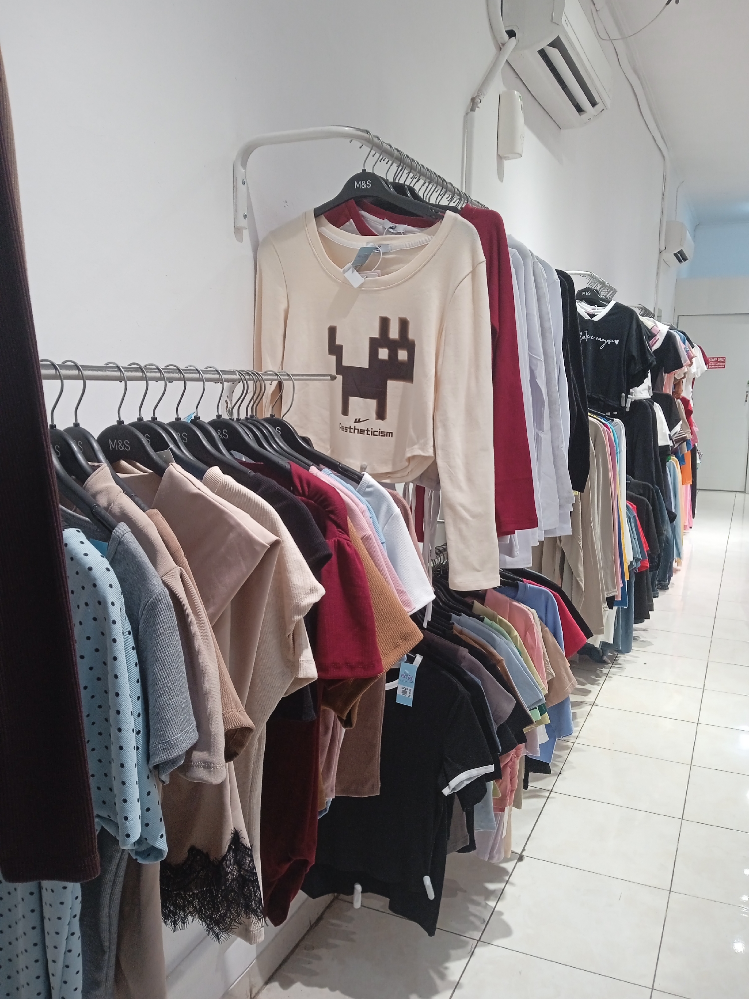 The image depicts a clothing store, likely in Semarang, Indonesia, featuring racks of women's apparel. The store appears to be well-lit with white tiled floors and white walls. Overhead, air conditioning units are visible. The clothing is organized on metal hangers and displayed on multiple clothing racks. Various styles and colors of tops, including t-shirts and long-sleeved shirts, are present. Some garments feature graphic prints, such as a pixelated black character on a cream-colored long-sleeved shirt. The hangers have "M&S" branding on some of them, suggesting a possible retail chain. The overall impression is of a typical retail environment for women's fashion.