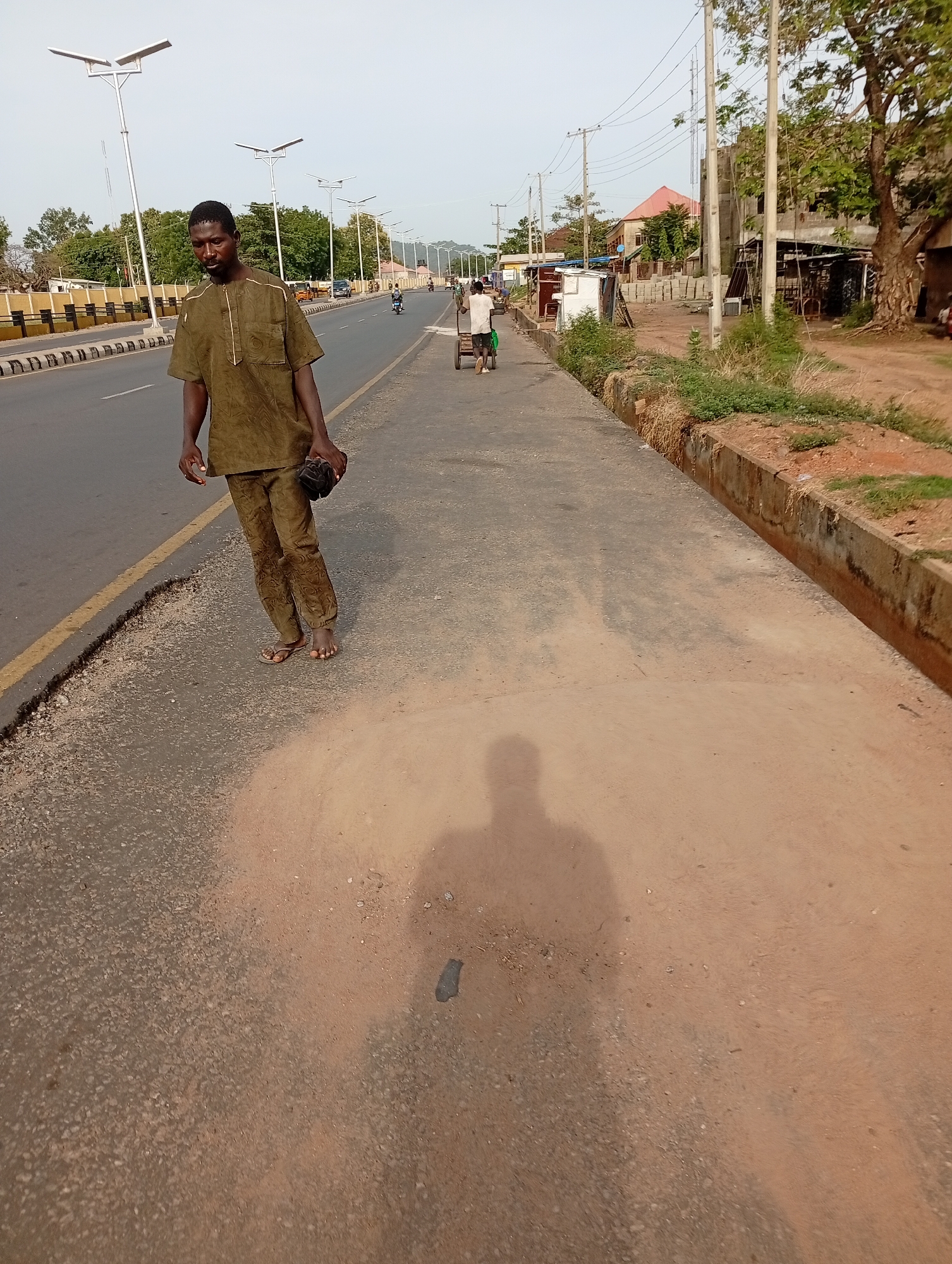 A man in brown clothing walks along a paved path adjacent to a road in Zango-ndaji, Nigeria. The path is partially covered in a reddish-brown soil or dust. The man is barefoot and carries a dark object in his left hand. His shadow is visible on the ground. A man pulling a cart is visible further down the path. Streetlights line the road. Buildings and trees are in the background. The roadside has a raised concrete edge with some vegetation growing along it.