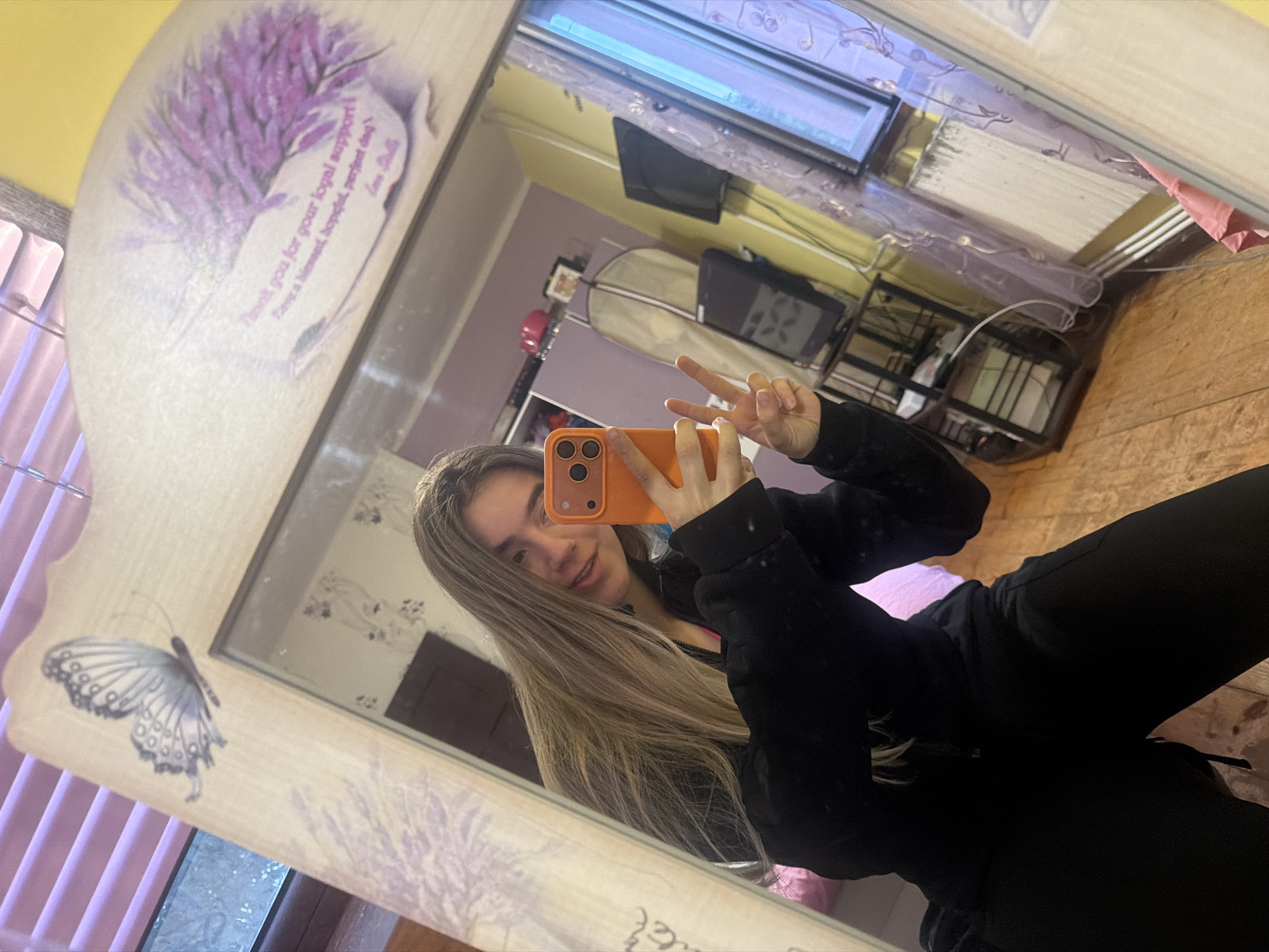 A young woman poses for a selfie in a mirror. She holds an orange cell phone, making a peace sign with her other hand. She has long, blonde hair and wears a black jacket. The mirror has a decorated frame with a butterfly and lavender design. The reflection shows a bedroom with a TV, dresser, and a window with blinds.
