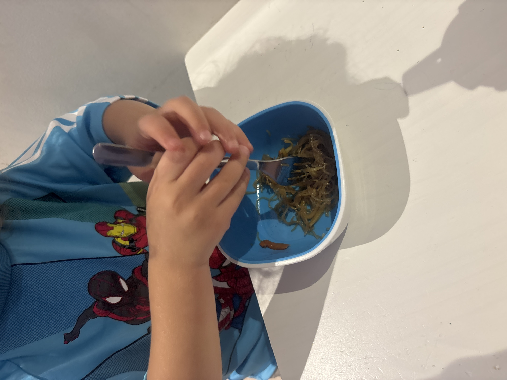 A close-up, eye-level shot shows a young person's hands using a fork to eat from a blue bowl. The person is wearing a blue long-sleeved shirt with graphic prints of Spider-Man and Iron Man. The bowl contains thin, tangled strands of food, possibly noodles or seaweed, with a dollop of dark sauce on the side. The table surface is white and appears to have some smudges. The background is a plain white wall. The image does not contain any information about the location being Dubai, United Arab Emirates.