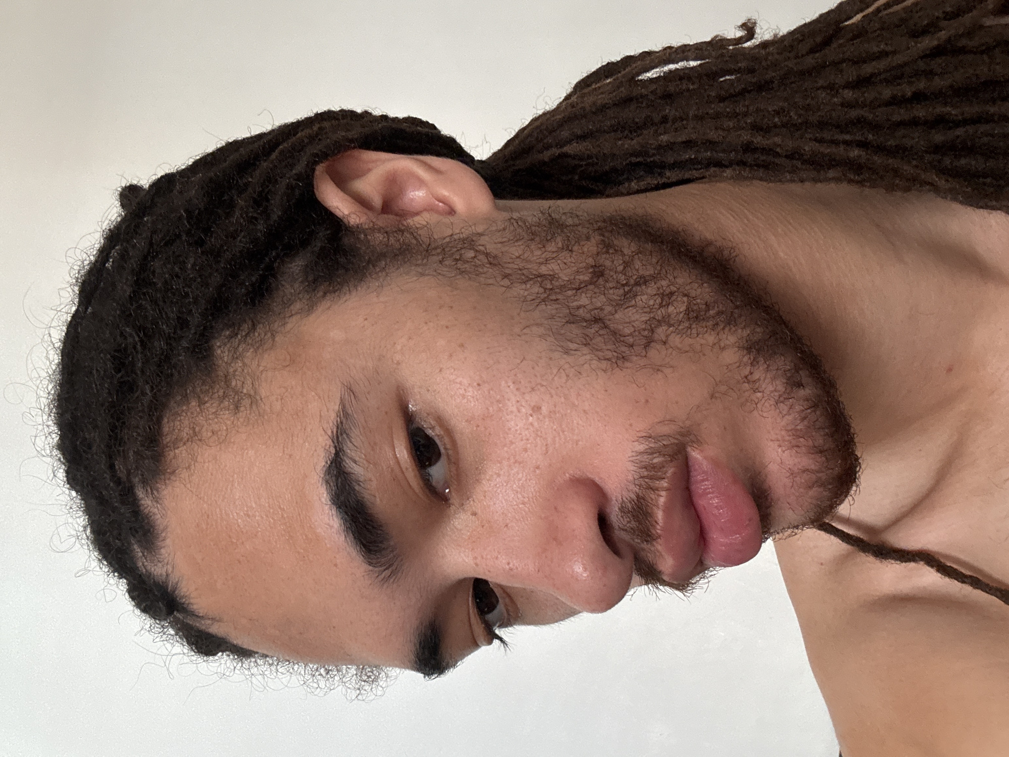 This is a close-up, head-and-shoulders portrait of a person with dark dreadlocks and a light beard. The subject is looking down and to the left, with their face angled away from the camera. Their skin is light brown with freckles, and they have dark eyebrows and eyes. The background is a plain, light-colored wall. There are no discernible landmarks or environmental clues that would indicate a specific location, including Paris, France.