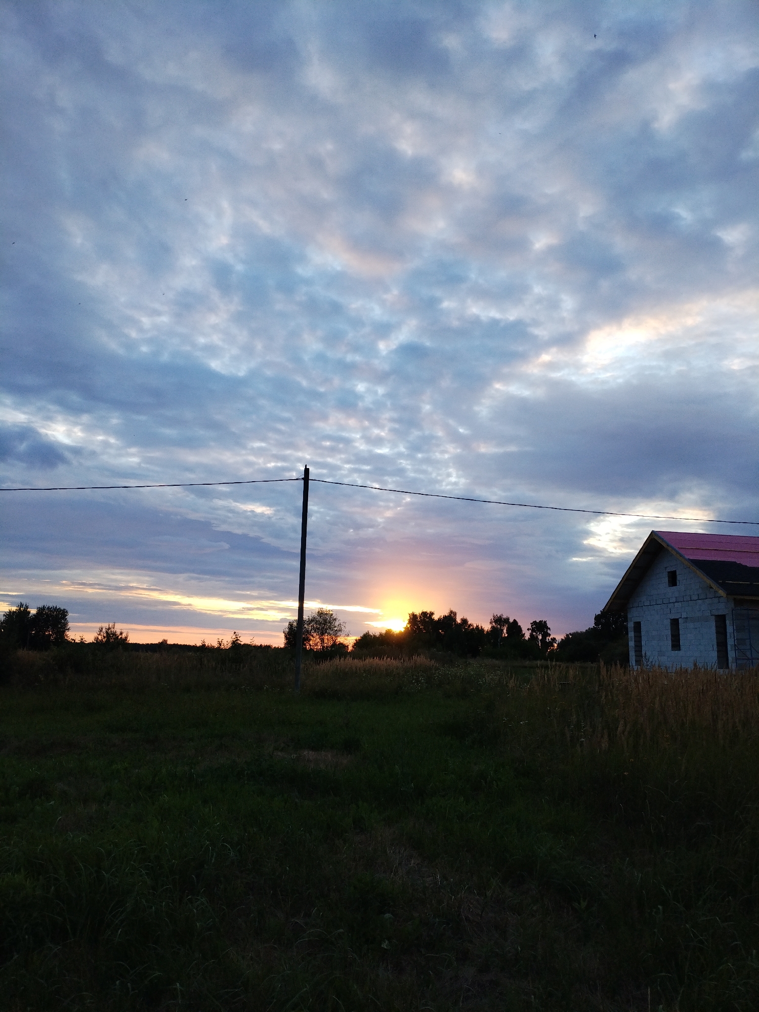 This is a landscape photograph taken during sunset in Bialynichy, Belarus. The foreground is dominated by a dark, grassy field. In the midground, a thin line of trees stretches across the horizon, partially obscuring the setting sun. The sun itself is a bright, orange orb, peeking through the foliage. To the right of the frame, a partially constructed house stands out against the dusky sky. It appears to be made of white bricks and has a pink roof. A single utility pole with a wire stretching across the frame is positioned in the center-left of the image. The sky is filled with dramatic clouds, tinged with shades of blue, gray, and a hint of pink from the sunset. A few small birds are visible in the sky. The overall impression is one of rural tranquility during twilight.