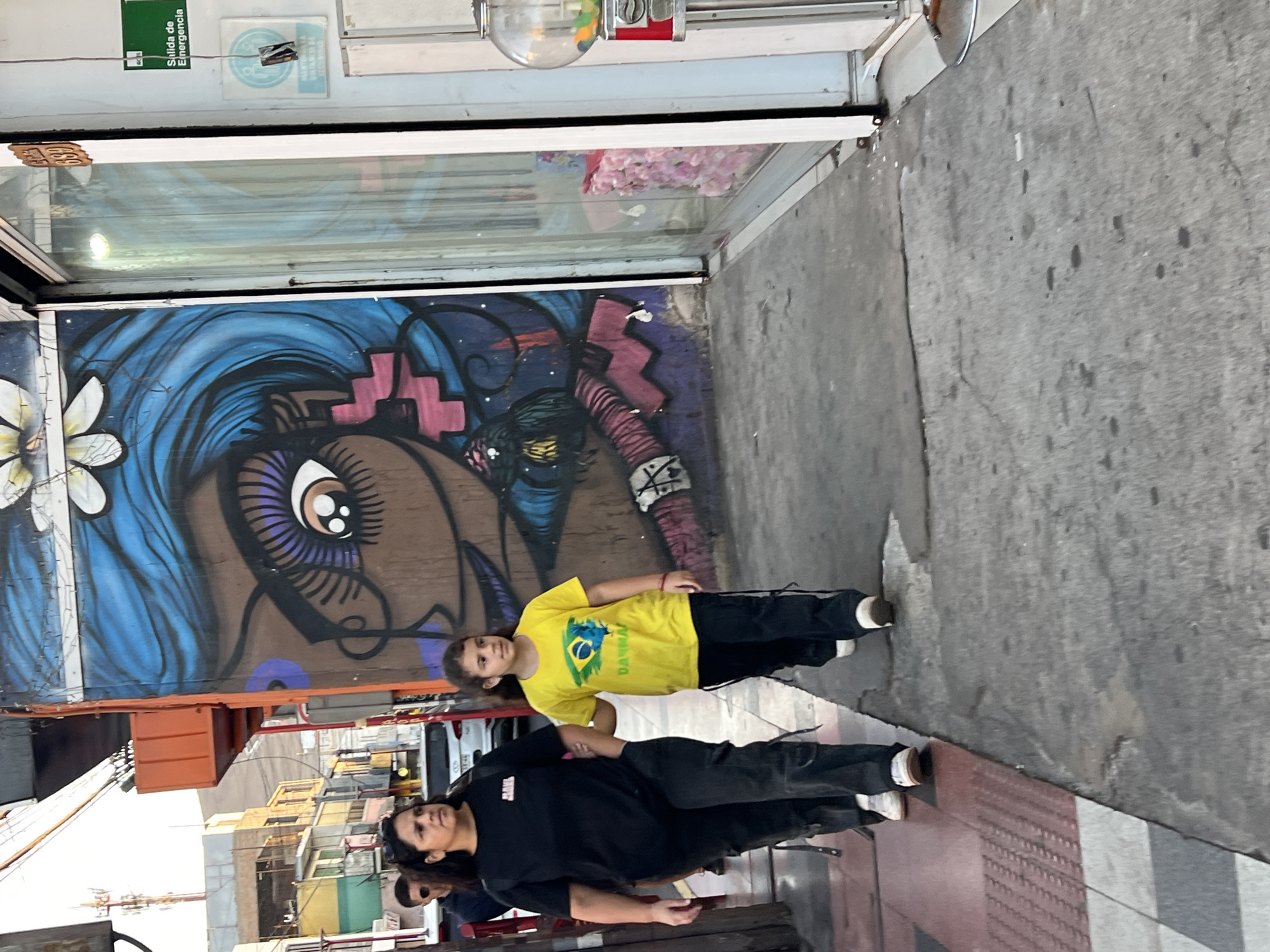 A street scene in Arica, Chile, depicts a mother and daughter standing beside a mural. The mural, painted on the exterior wall of a building, features a large eye with blue hair and purple accents. The mother is wearing black clothing, and the daughter is wearing a yellow t-shirt with the Brazilian flag. They are standing on a grey sidewalk with a red and grey tiled border. The background shows other buildings and urban elements.