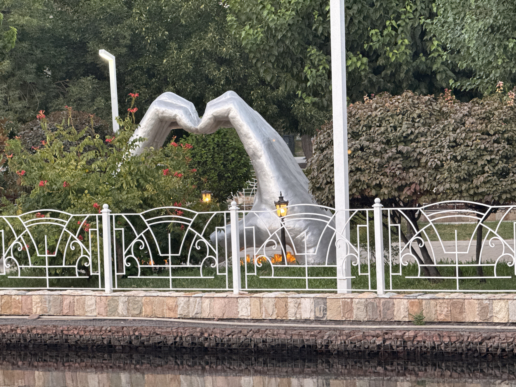 The image shows a large, metallic sculpture of two hands forming a heart shape, positioned behind a white decorative fence. The sculpture is silver and appears to be made of polished metal. Behind the sculpture and fence, there are lush green trees and bushes. Several small, yellow street lamps are visible, some illuminating the sculpture and others the surrounding foliage. The fence is ornate with geometric patterns and curves. In front of the fence, there is a stone wall with varying colors of stone, and below that, a gabion wall filled with rocks. A body of dark water, likely a canal or river, is in the foreground, reflecting the scene above. The lighting suggests it is either early morning or late evening. This sculpture is located in Tashkent, Uzbekistan, specifically in the Magic City amusement park. There are no people visible in the image.