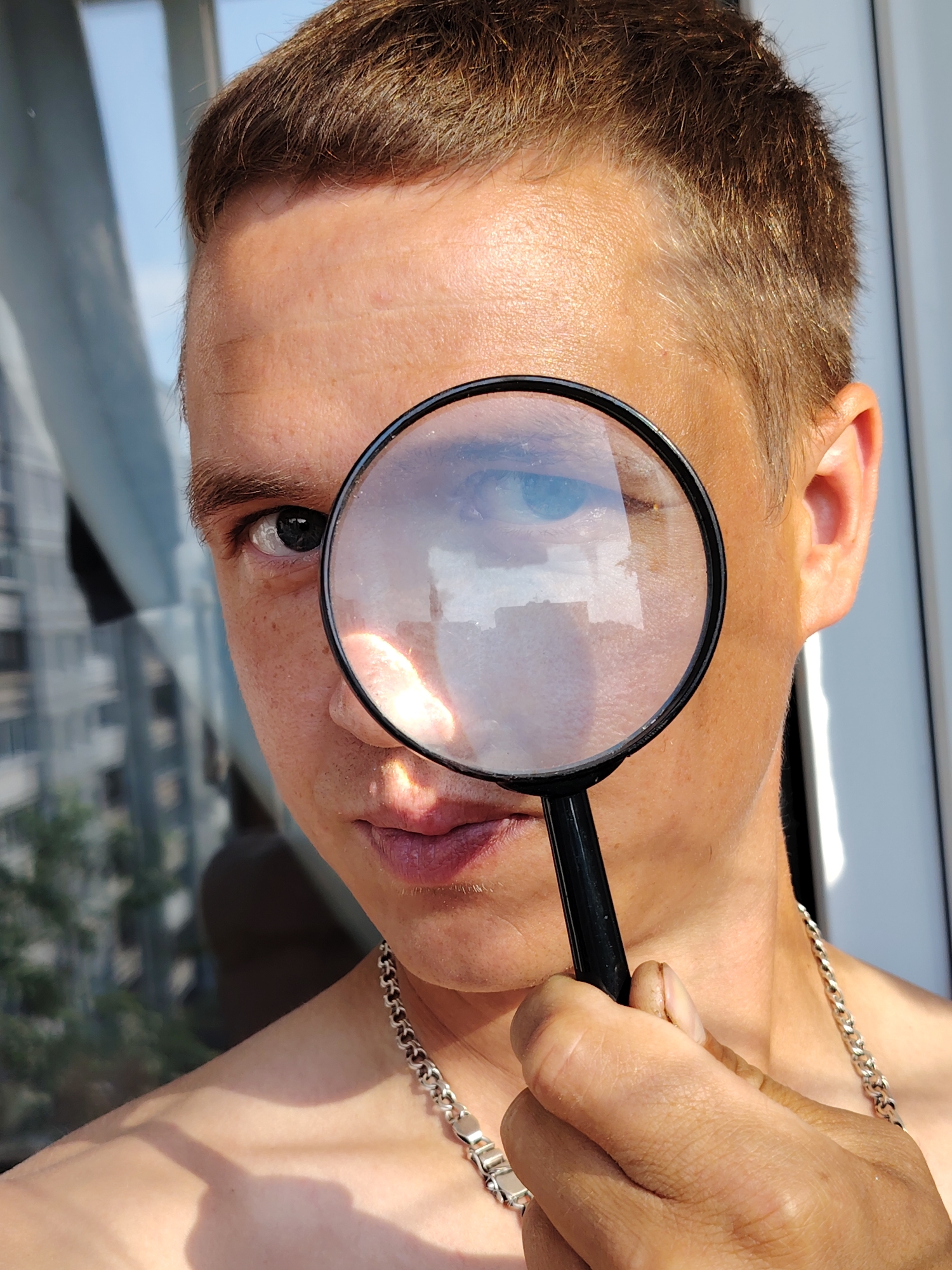 A close-up, eye-level, outdoor shot features a person holding a magnifying glass to their left eye. The subject's head and shoulders are visible, with their hair a light brown. The magnifying glass is black-rimmed and held by a black handle. The person is wearing a silver chain necklace. The background is blurred, showing elements of a city, possibly Chaikovsky, Russia, with buildings and a bright sky. The lighting suggests a sunny day.