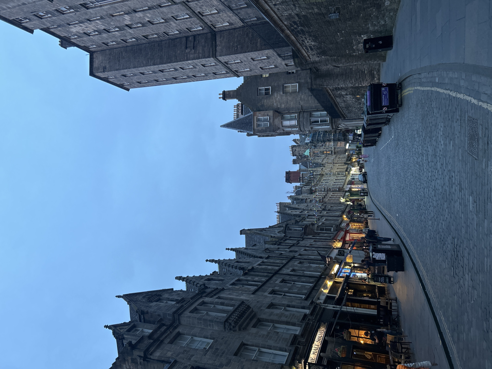 The image depicts a cobblestone street in the Old Town of Edinburgh, Scotland, during twilight. The street is lined with historic stone buildings, many with steeply pitched roofs and ornate architectural details. Several shops and businesses are visible along the left side of the street, with illuminated signs and window displays. A few people are walking on the pavement and a restaurant appears to have outdoor seating. On the right side of the street, a tall stone building dominates the foreground, with a series of large black bins lined up along the pavement. The sky is a pale blue, indicating the transition from day to night.