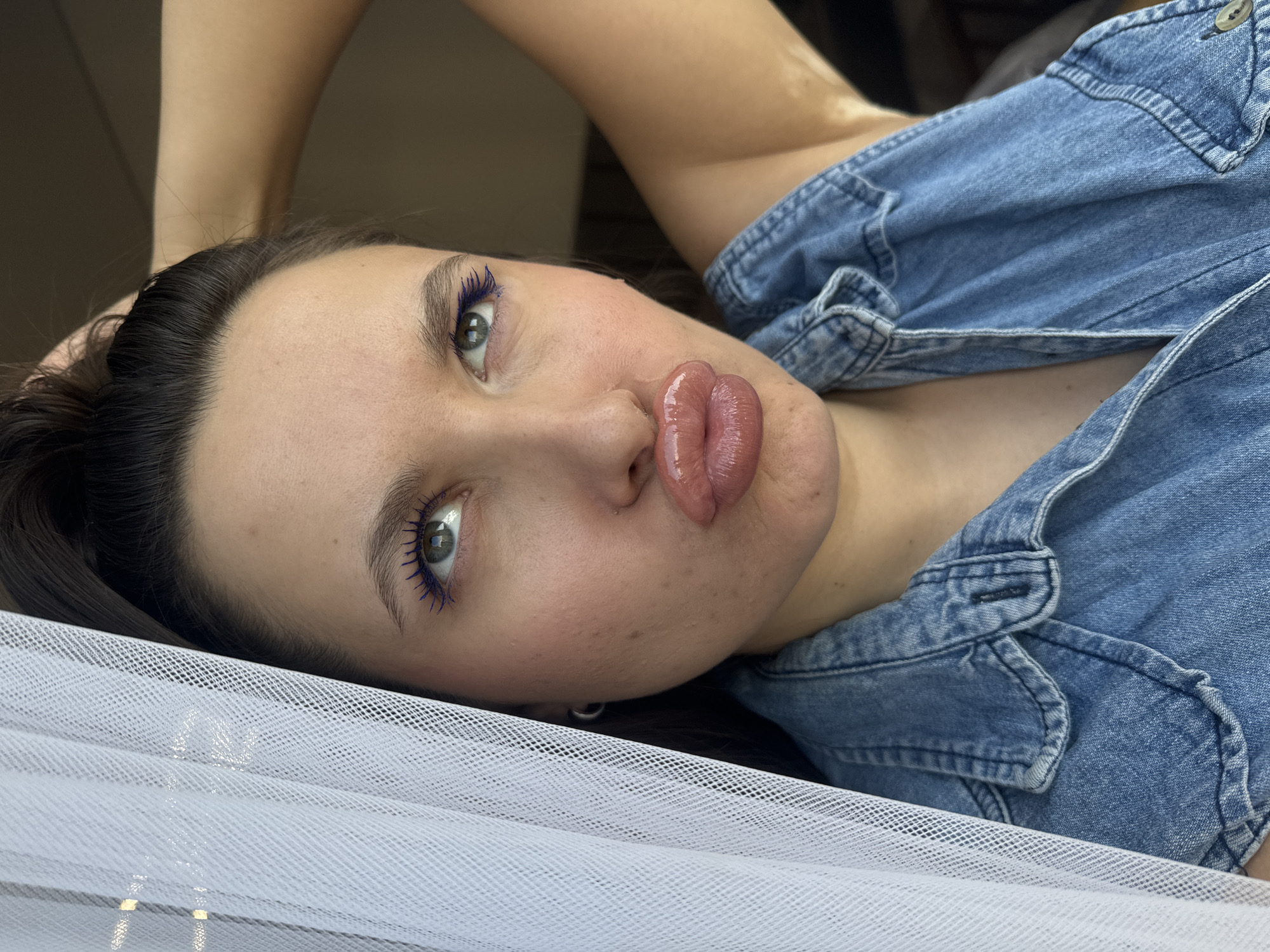 A woman with dark hair is lying down, her head resting on a white, net-like fabric. She is wearing a blue denim shirt. She has blue mascara on her eyelashes. The location, Akulovo (Gp Kubinka), Russia, is not determinable from the image.