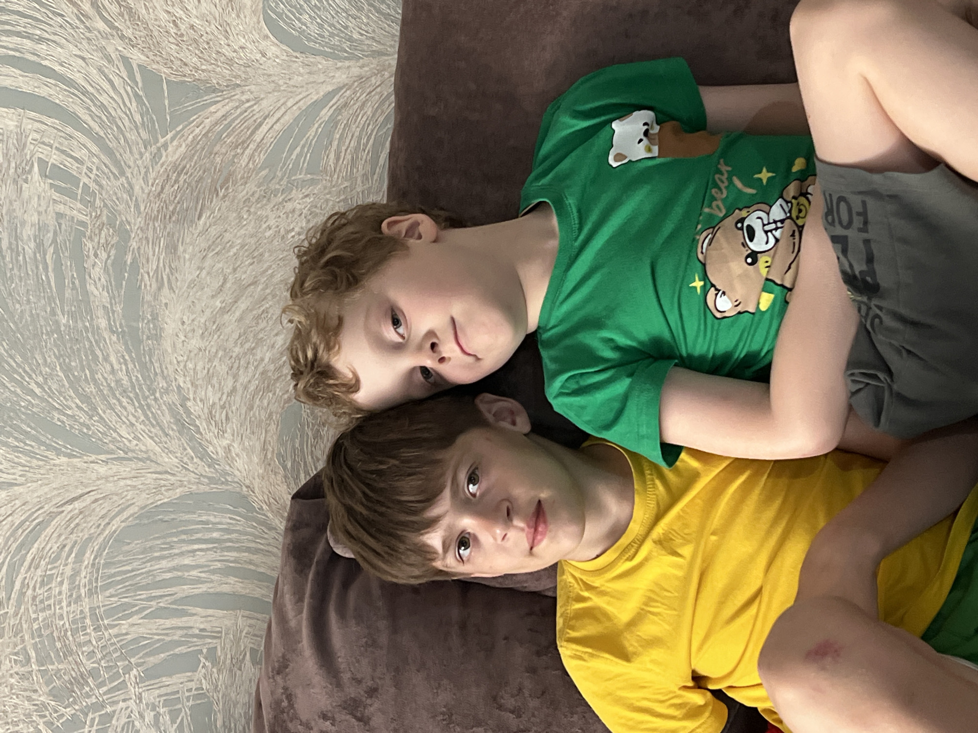 Two boys are lying on a brown couch in a room with patterned wallpaper. The first boy, with curly blonde hair, is wearing a green t-shirt with a bear design and grey shorts. The second boy, with short brown hair, is wearing a yellow t-shirt and green shorts. They are positioned closely together, looking towards the camera. The location in the city of Noviy, Russia cannot be determined from the image.