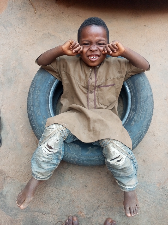 The image portrays a young child seated within a black rubber vehicle tire on an unpaved dirt ground in Tempure, Nigeria. The child, identified as a boy, has dark skin and short, dark hair. He wears a light brown, long-sleeved tunic with dark brown embroidered details on the placket and a chest pocket, over light blue distressed denim jeans with visible tears on both knees. His feet are bare and rest on the ground in front of the tire. He faces the camera directly, displaying an exaggerated, wide-grinning expression with squinted eyes and an open mouth revealing teeth. His hands are raised to the sides of his face, fingers curled into loose fists near his cheeks. The tire's sidewall shows markings including "GOODRIDE RADIAL" and partial size information. A light brown or beige wall forms the background. Two bare adult feet are partially visible on the ground at the bottom center of the frame.
